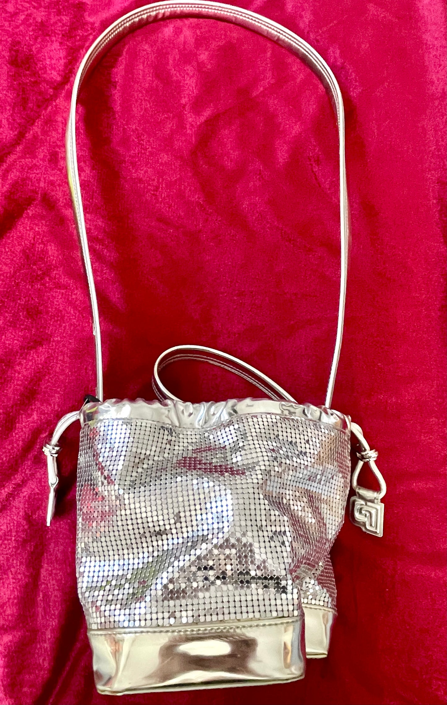 Full view of Paco Rabanne silver shoulder/wrist bag with long strap shown, on red velvet fabric background.