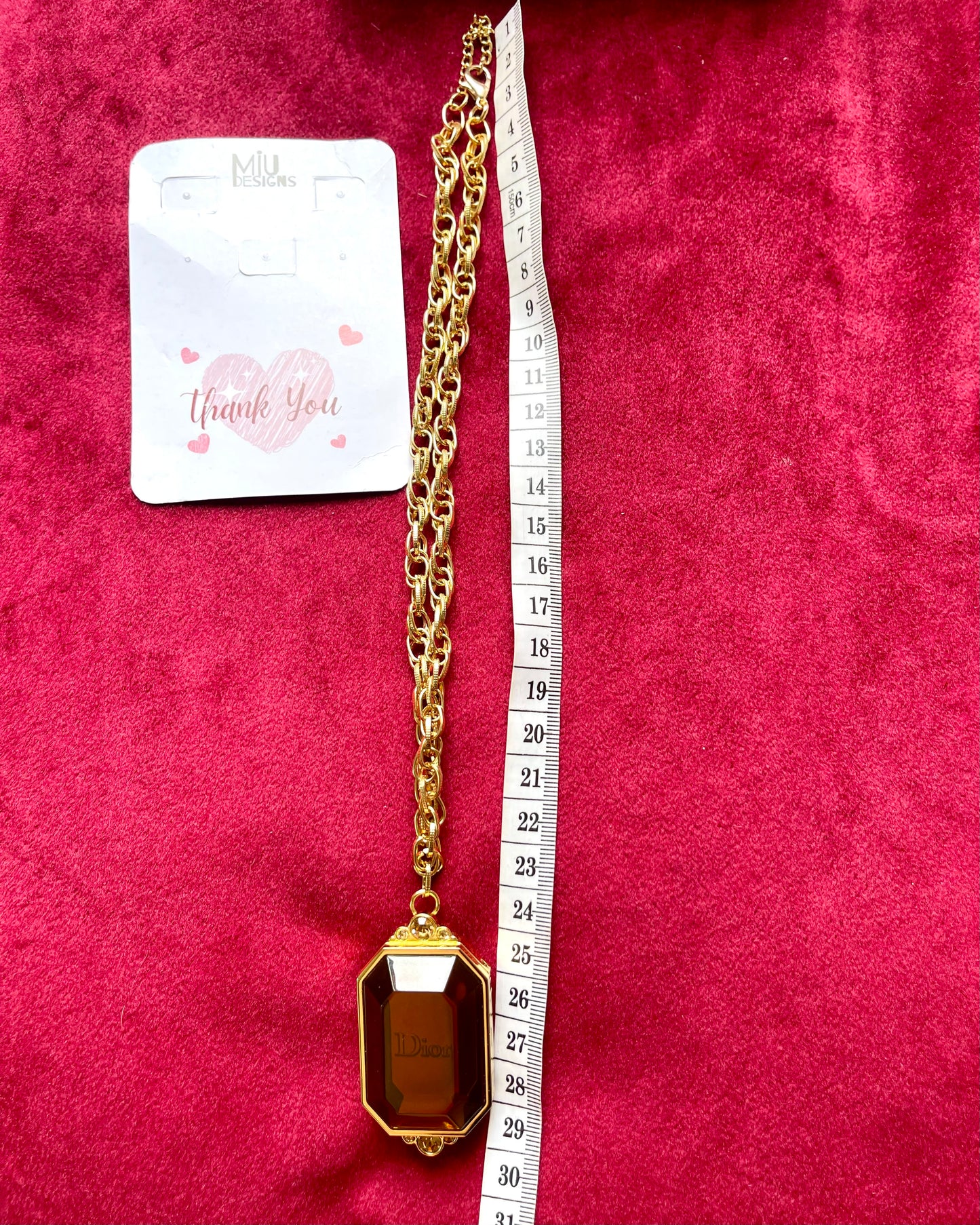 Golden vintage Dior couture pendant locket necklace with amber gem and Dior engraving on red velvet background using measuring tape showcasing Miu Designs brand for necklace
