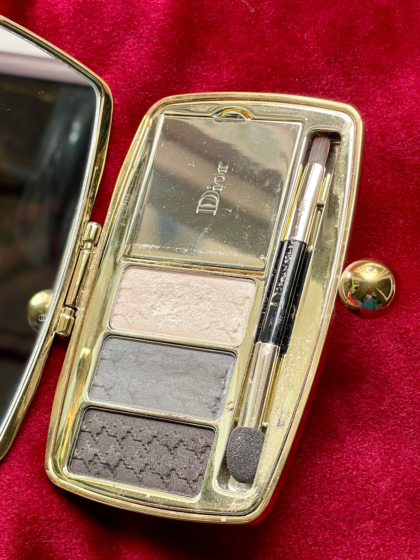 Close up Open vintage Dior Minaudière makeup palette showing mirror, three eyeshadows in neutral shades, and dual-ended applicators inside a gold-trimmed compact on a red velvet background