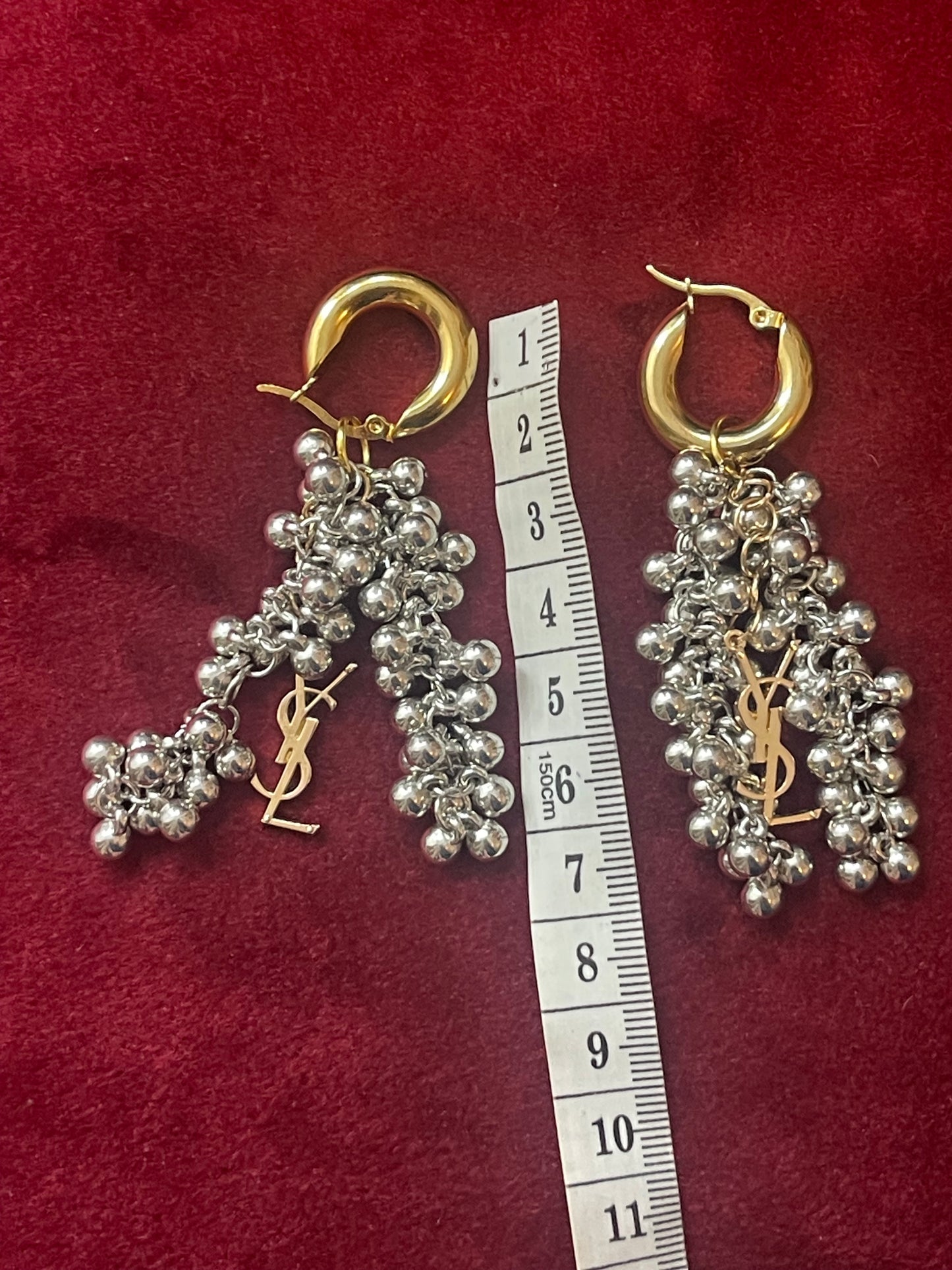 Pair of silver beaded earrings with gold Yves Saint Laurent logo on a red background with a ruler for scale.