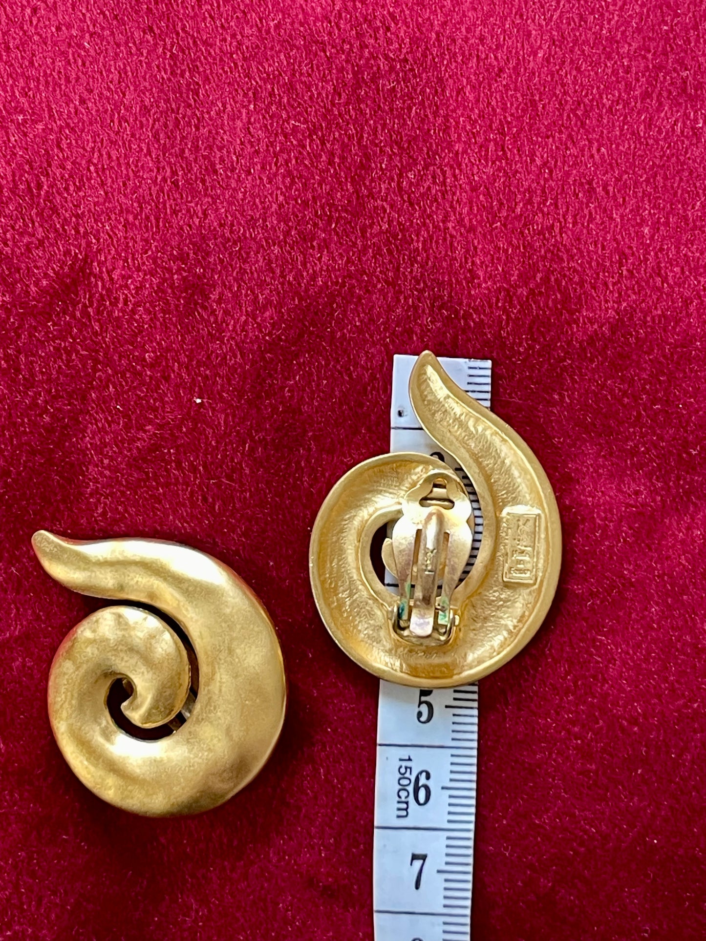 1980s Yves Saint Laurent gold tone spiral clip-on earrings with designer stamp, shown on red fabric background with a measuring tape