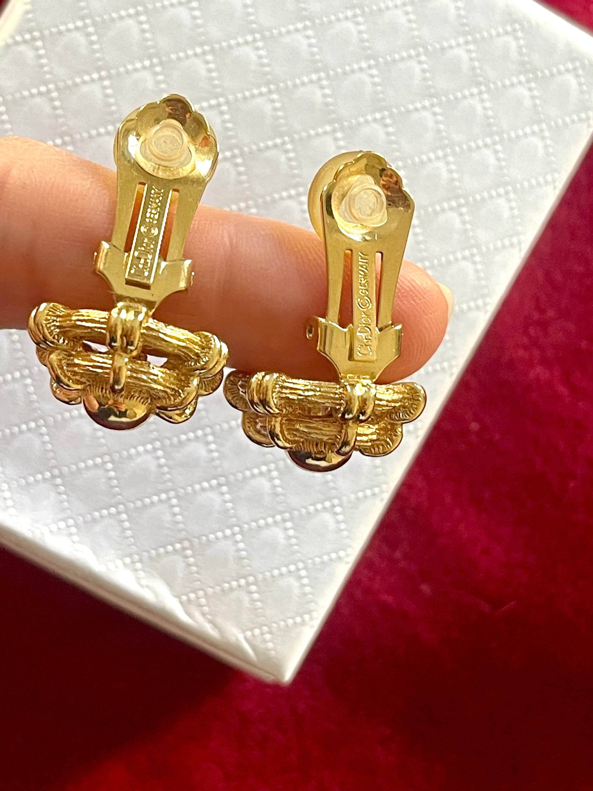 Dior Gold earrings held between fingers showing the signature against a white box background