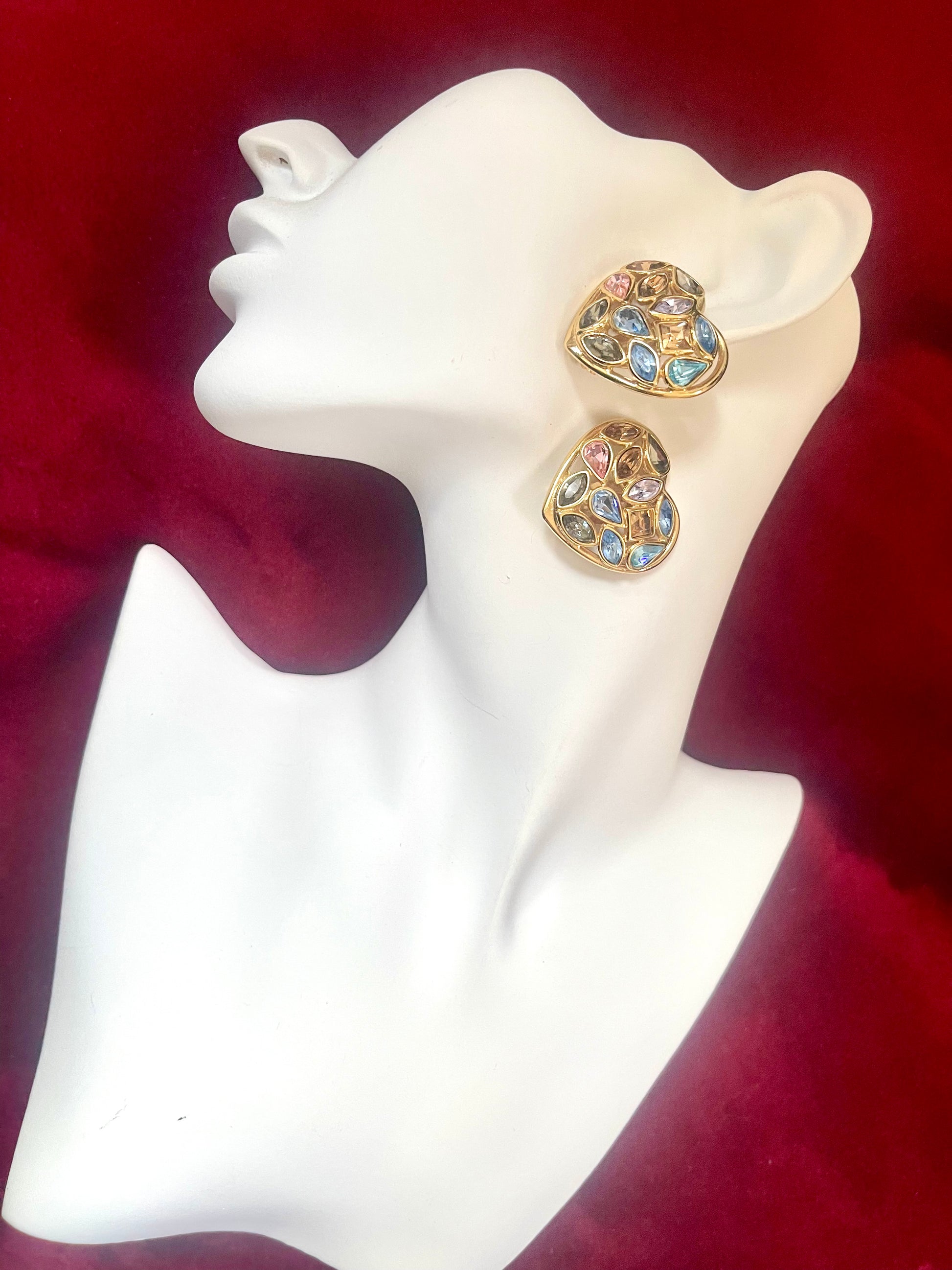 Pair of gold-tone vintage Yves Saint Laurent heart-shaped clip-on earrings decorated with multi-colored crystals, displayed on a mannequin bust on a red background