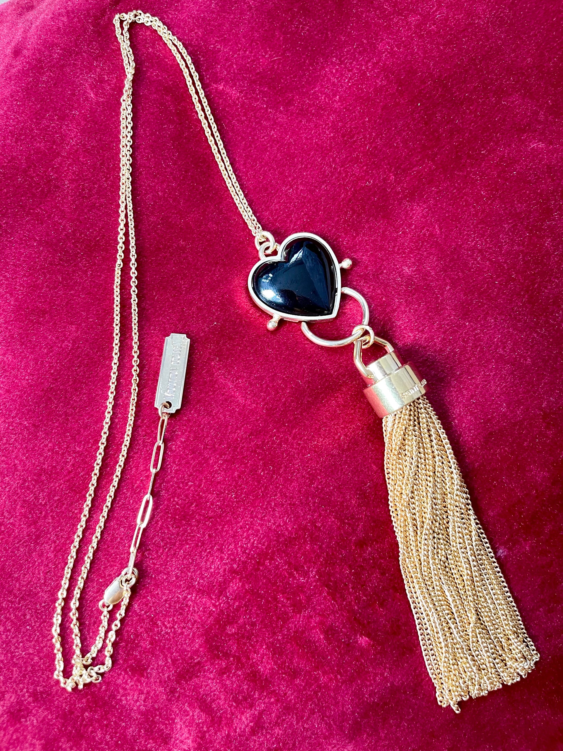 Gold-plated vintage necklace with black resin heart pendant and attached Yves Saint Laurent gold tassel charm displayed on red velvet.