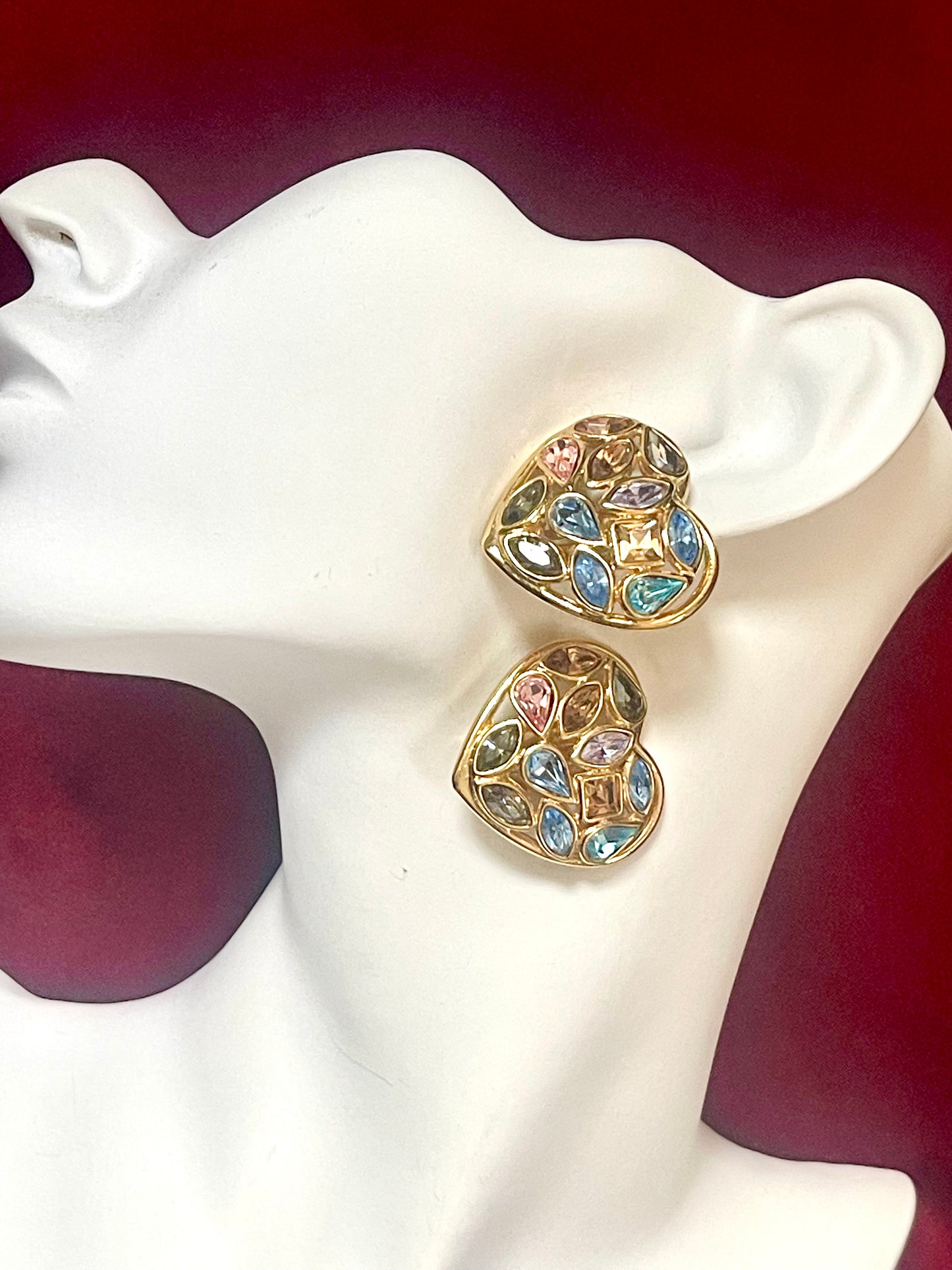 Pair  gold-tone vintage Yves Saint Laurent heart-shaped clip-on earrings decorated with multi-colored crystals, displayed on a mannequin on a red background