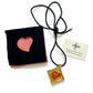 Gold heart-shaped pendant on a black cord with a black pouch featuring a pink heart and a card labeled 'Yves Saint Laurent' on a white background.