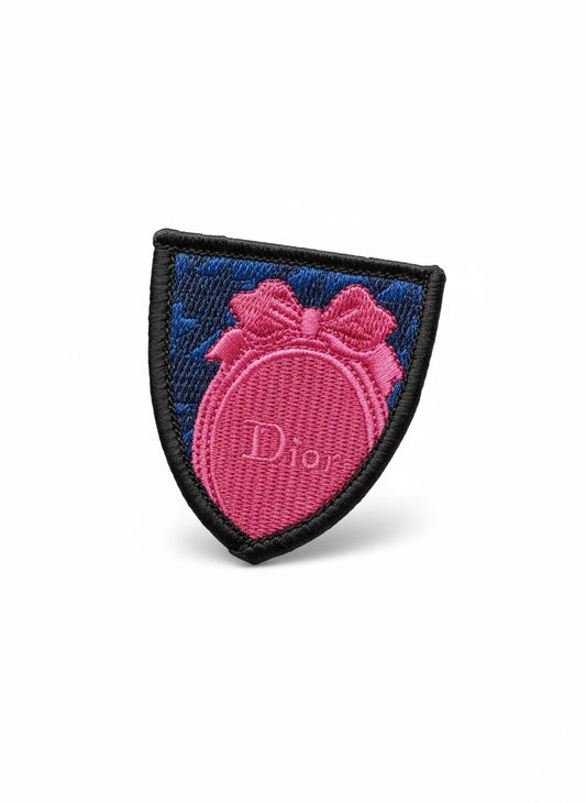 A vintage Dior embroidered brooch with a metal aluminum base featuring pink and blue colors and the Dior logo on white background 