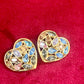Pair of 3 cm gold-tone vintage Yves Saint Laurent heart-shaped clip-on earrings decorated with multi-coloured crystals, displayed on a red background