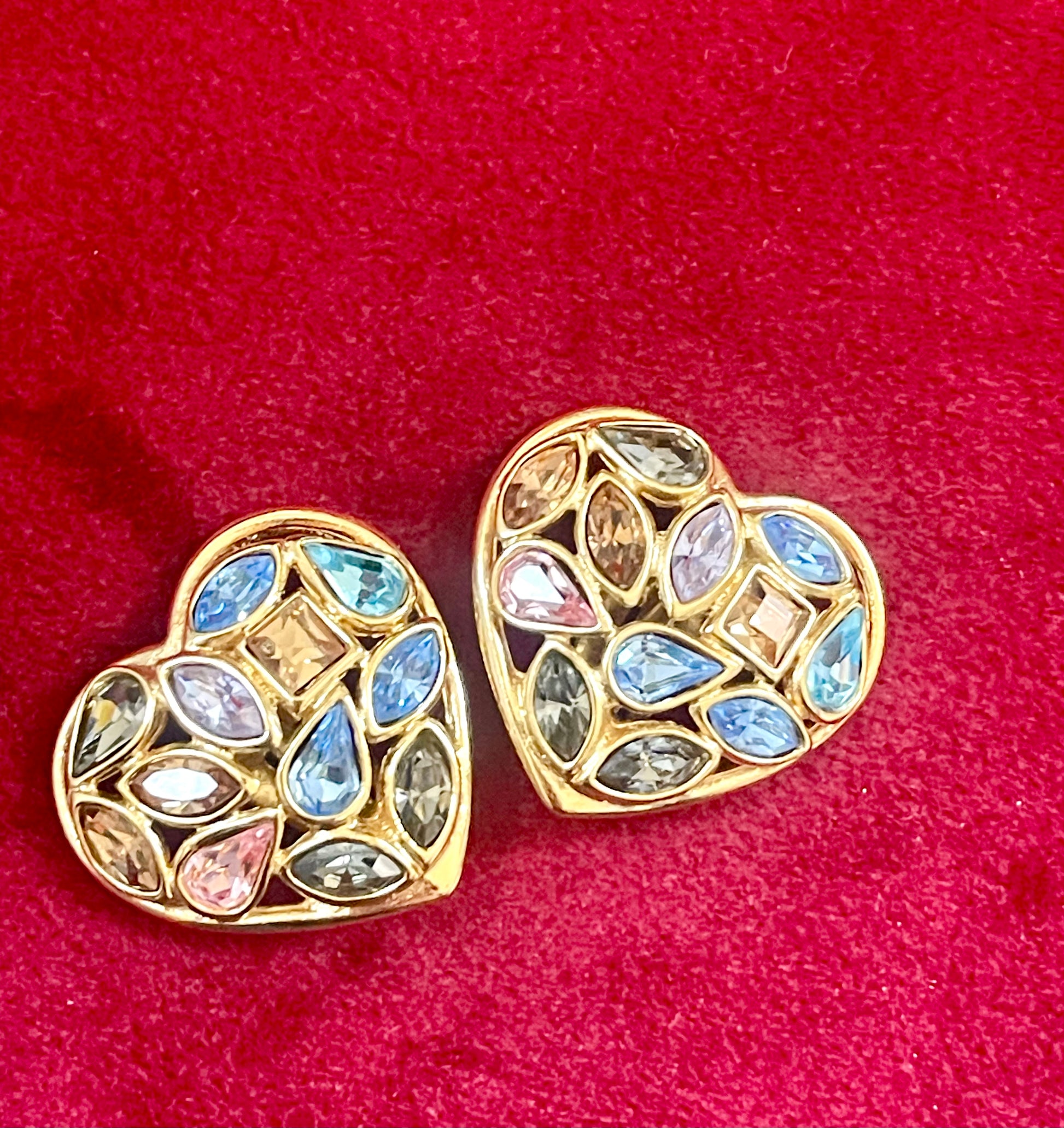 Pair of 3 cm gold-tone vintage Yves Saint Laurent heart-shaped clip-on earrings decorated with multi-coloured crystals, displayed on a red background