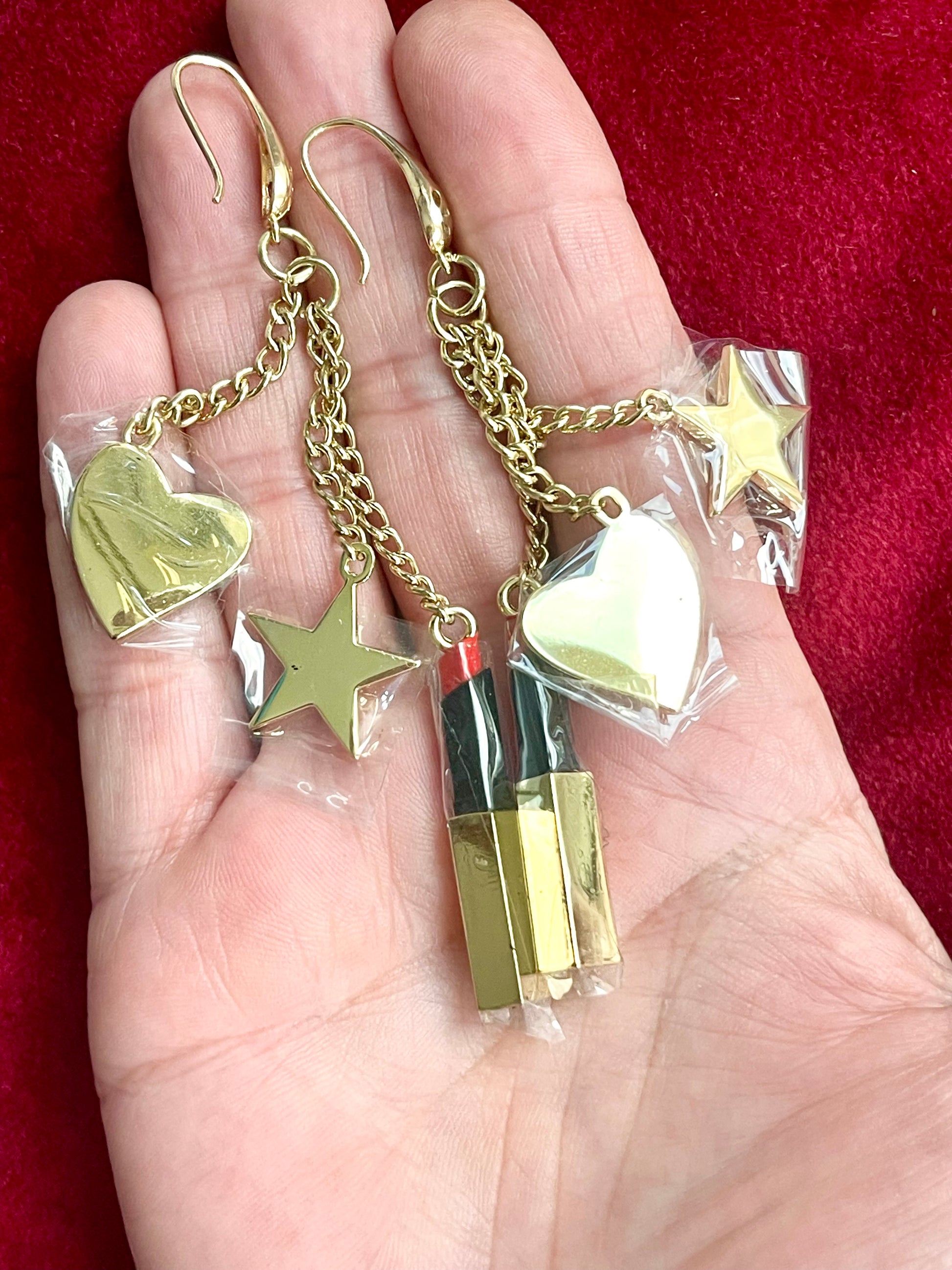 Back view YSL Gold earrings with heart, star, and lipstick charms held in a hand against a red background