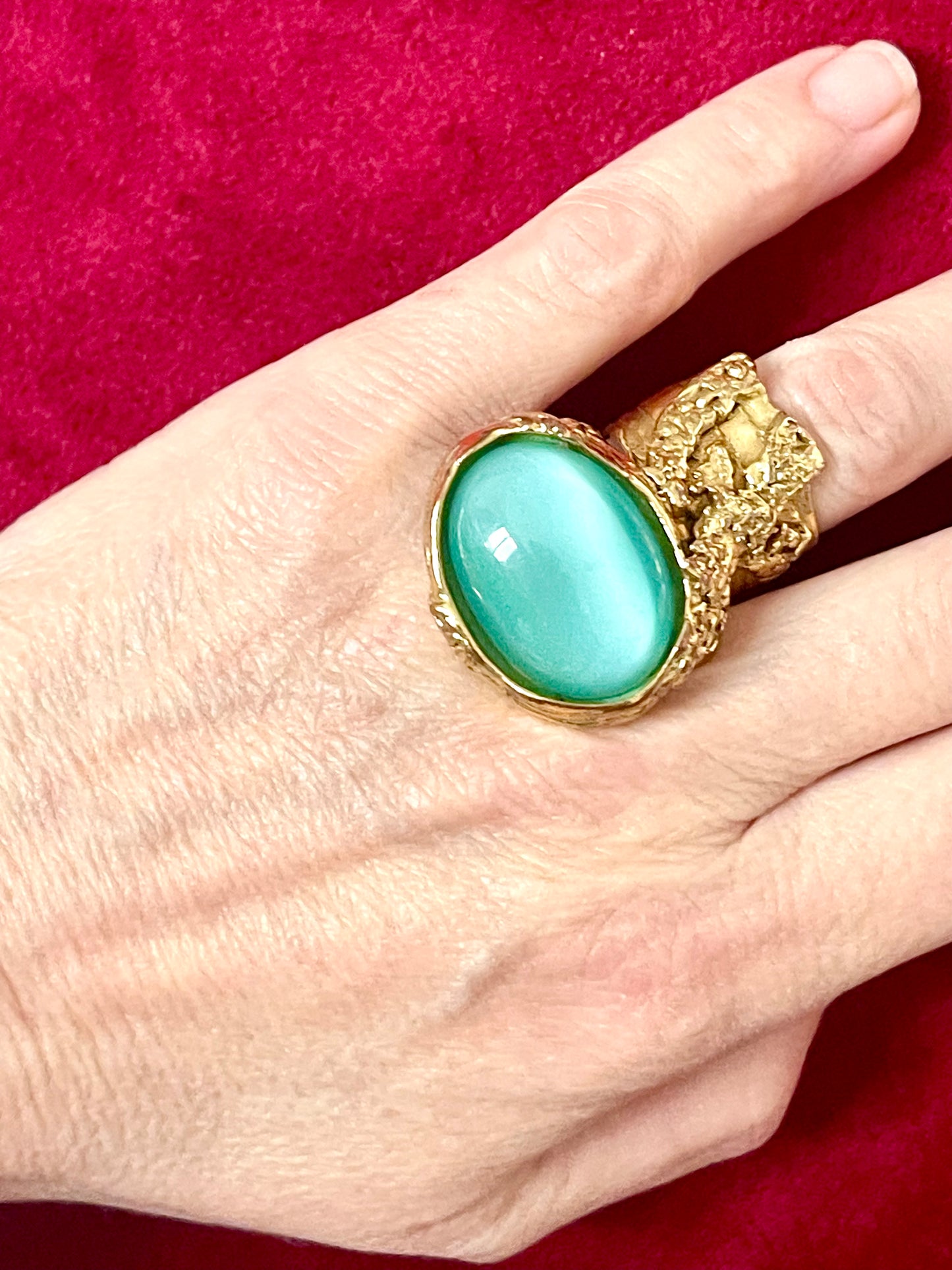 Hand wearing a bold vintage Yves Saint Laurent ring featuring a large turquoise nacre stone set in textured gold metal, photographed against a deep red velvet background.