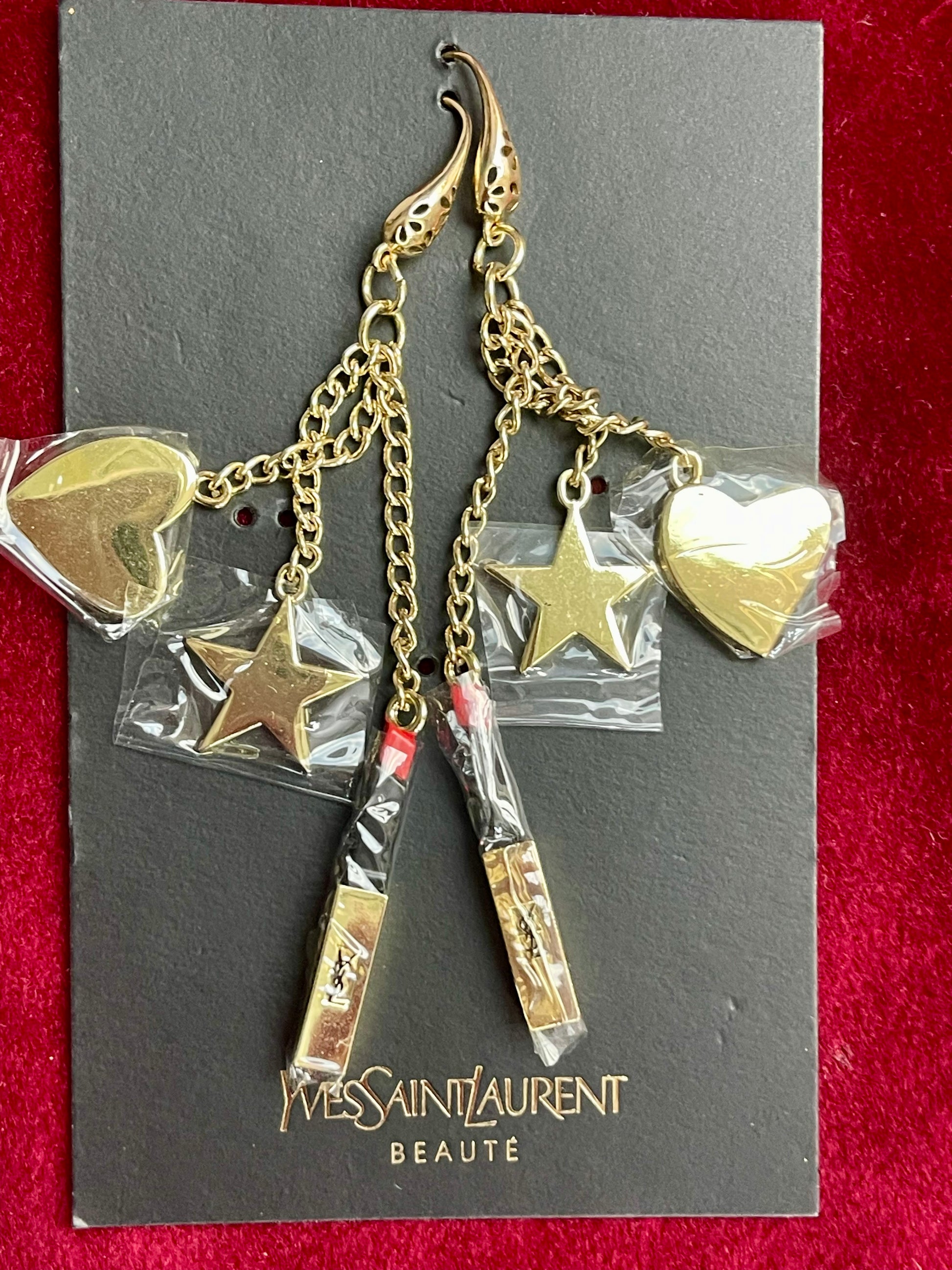 Gold earrings with heart and star charms on a Yves Saint Laurent card against a red background