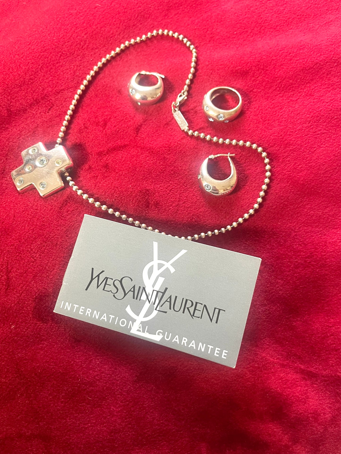 Vintage YSL silver jewelry set displayed on red fabric: silver earrings, cross pendant necklace on beaded chain, ring and YSL guarantee card