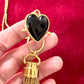 Close-up of black resin heart pendant paired with Yves Saint Laurent gold chain tassel charm held by hand on a red velvet background