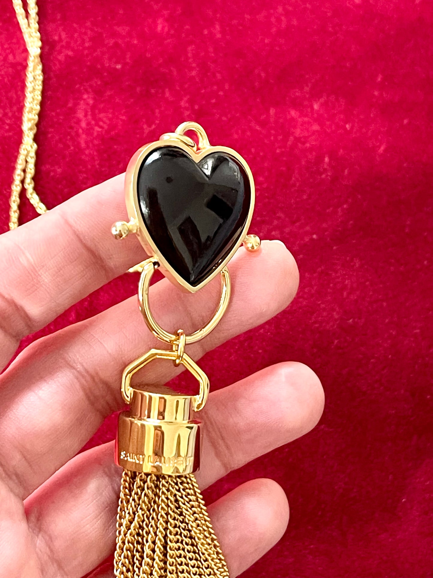 Close-up of black resin heart pendant paired with Yves Saint Laurent gold chain tassel charm held by hand on a red velvet background