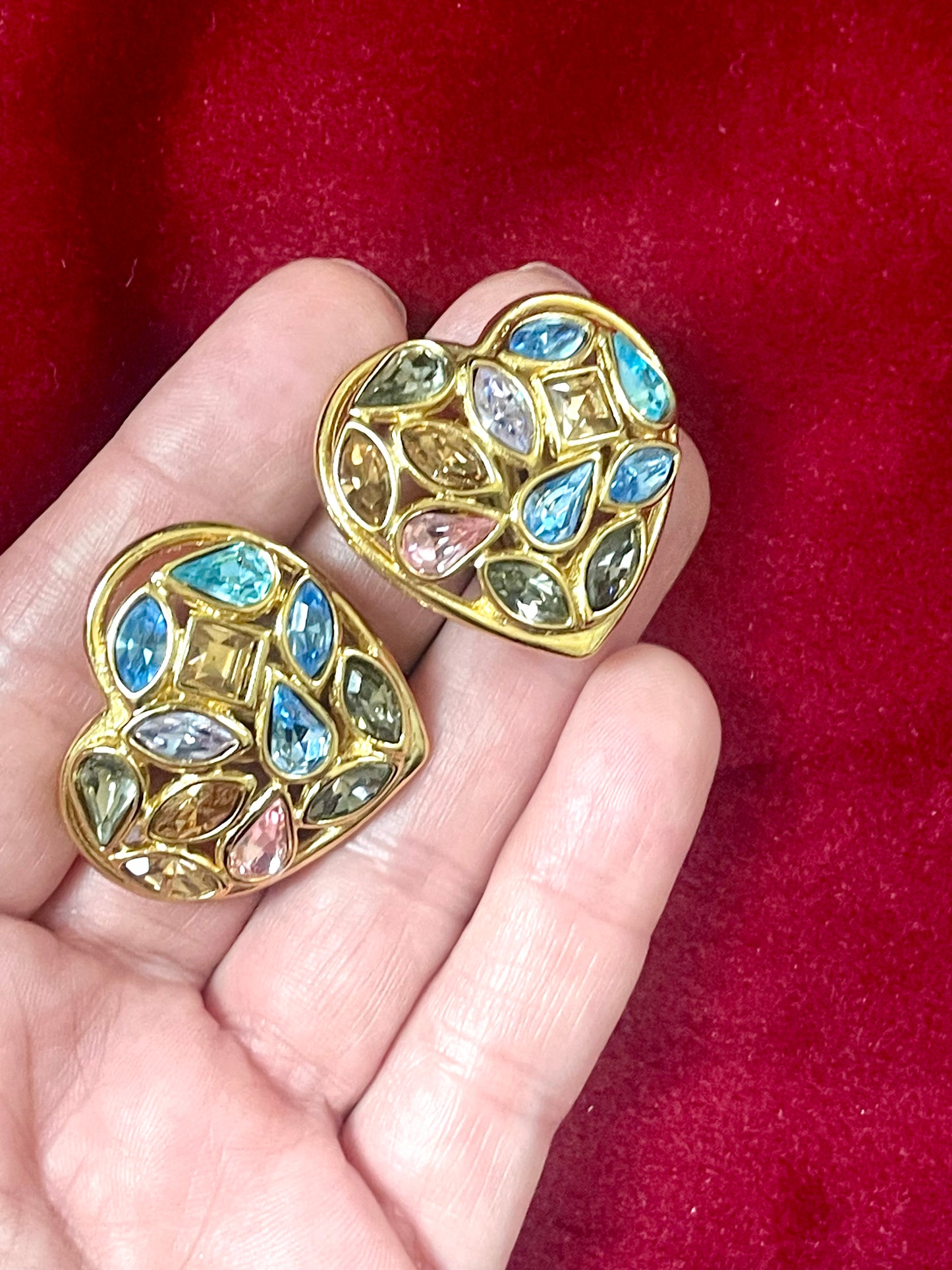 On hand Pair of 3 cm gold-tone vintage Yves Saint Laurent heart-shaped clip-on earrings decorated with multi-colored crystals, displayed on a red background