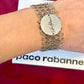 Silver bracelet with a circular tag on a wrist, against a red background with 'Paco Rabanne' branding.