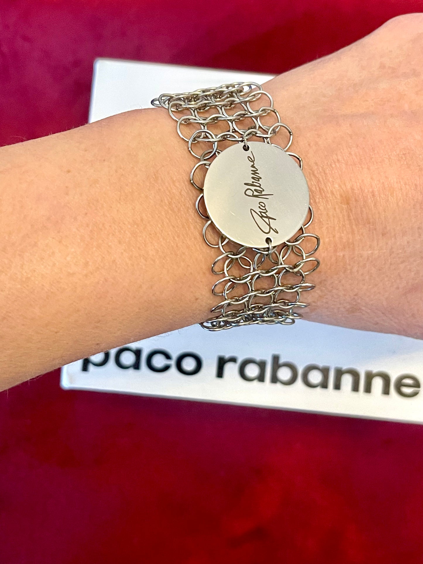 Silver bracelet with a circular tag on a wrist, against a red background with 'Paco Rabanne' branding.