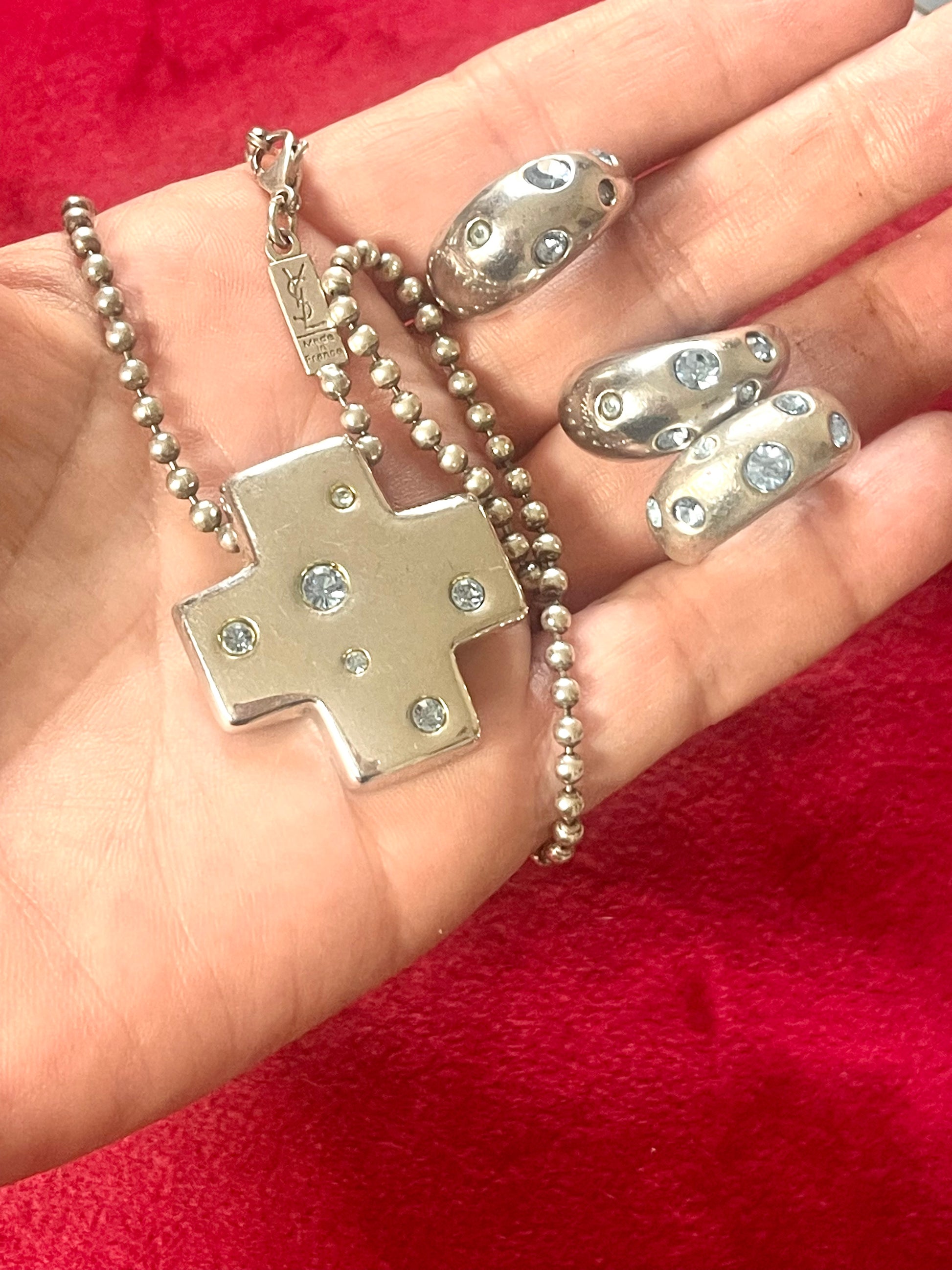 Vintage Yves Saint Laurent solid silver jewelry set displayed on red fabric: silver earrings, cross pendant necklace on beaded chain, ring displayed on hand 