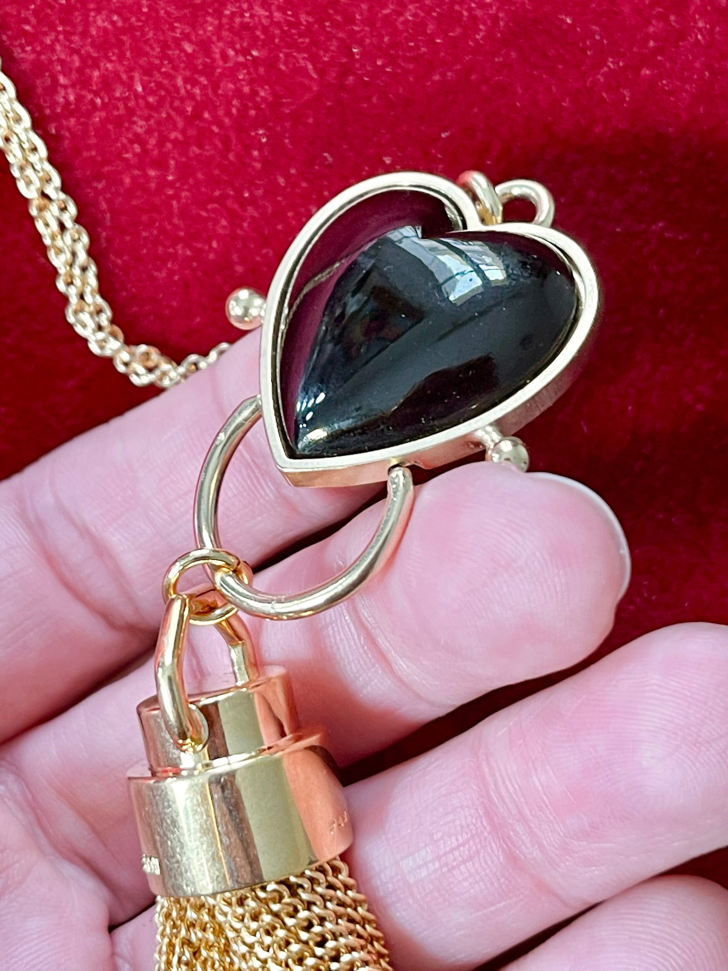 Close-up of black resin heart pendant paired with Yves Saint Laurent gold chain tassel charm held by hand on a red velvet background