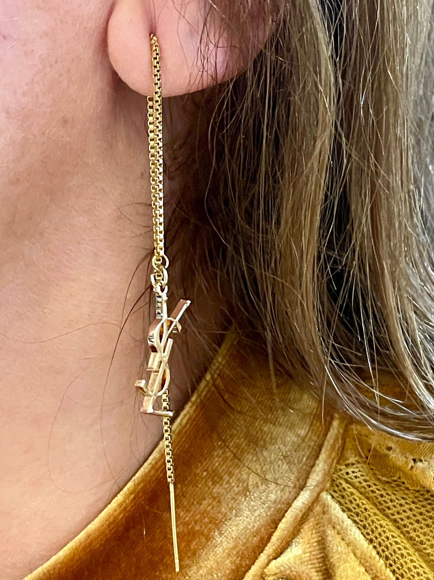 Gold earring with a small YSL pendant worn by a person with blonde hair.