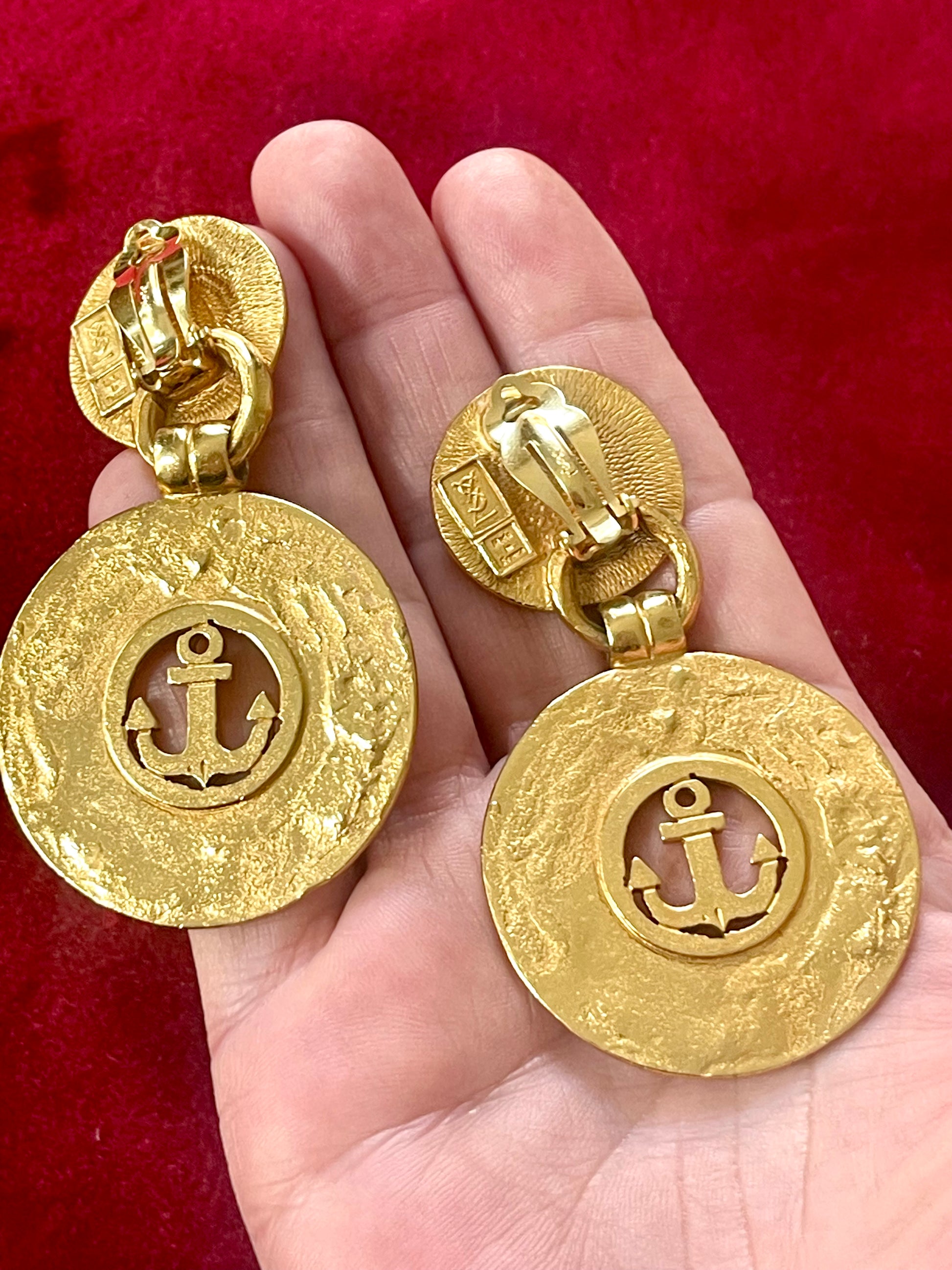 Back of Yves Saint Laurent gold-tone anchor earrings with engraved signatures red background Displayed on hand
