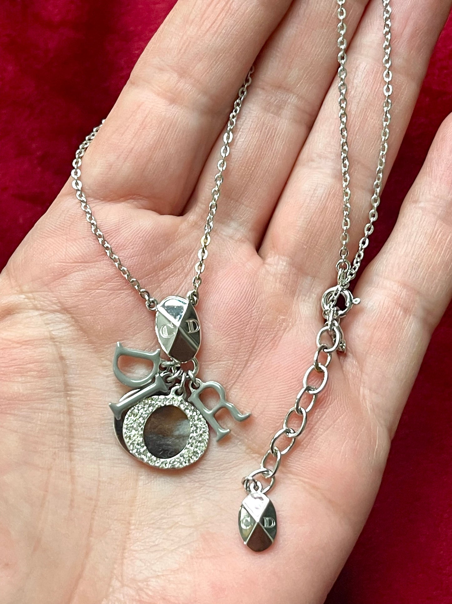 Short silver-tone Dior necklace with grey enameled D I O R letter charms and Dior oval tag, displayed on a hand against a red velvet background