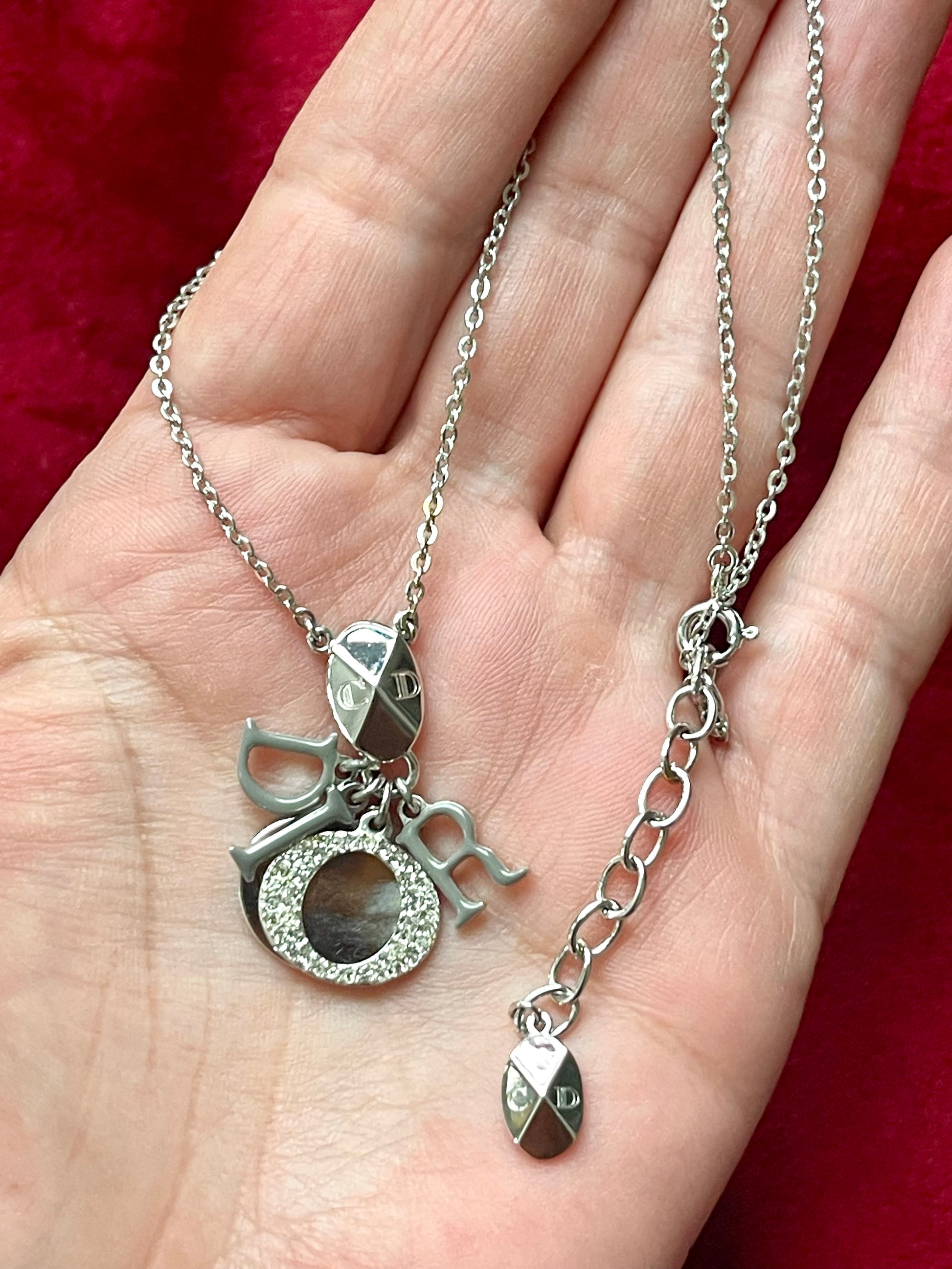 Short silver-tone Dior necklace with grey enameled D I O R letter charms and Dior oval tag, displayed on a hand against a red velvet background