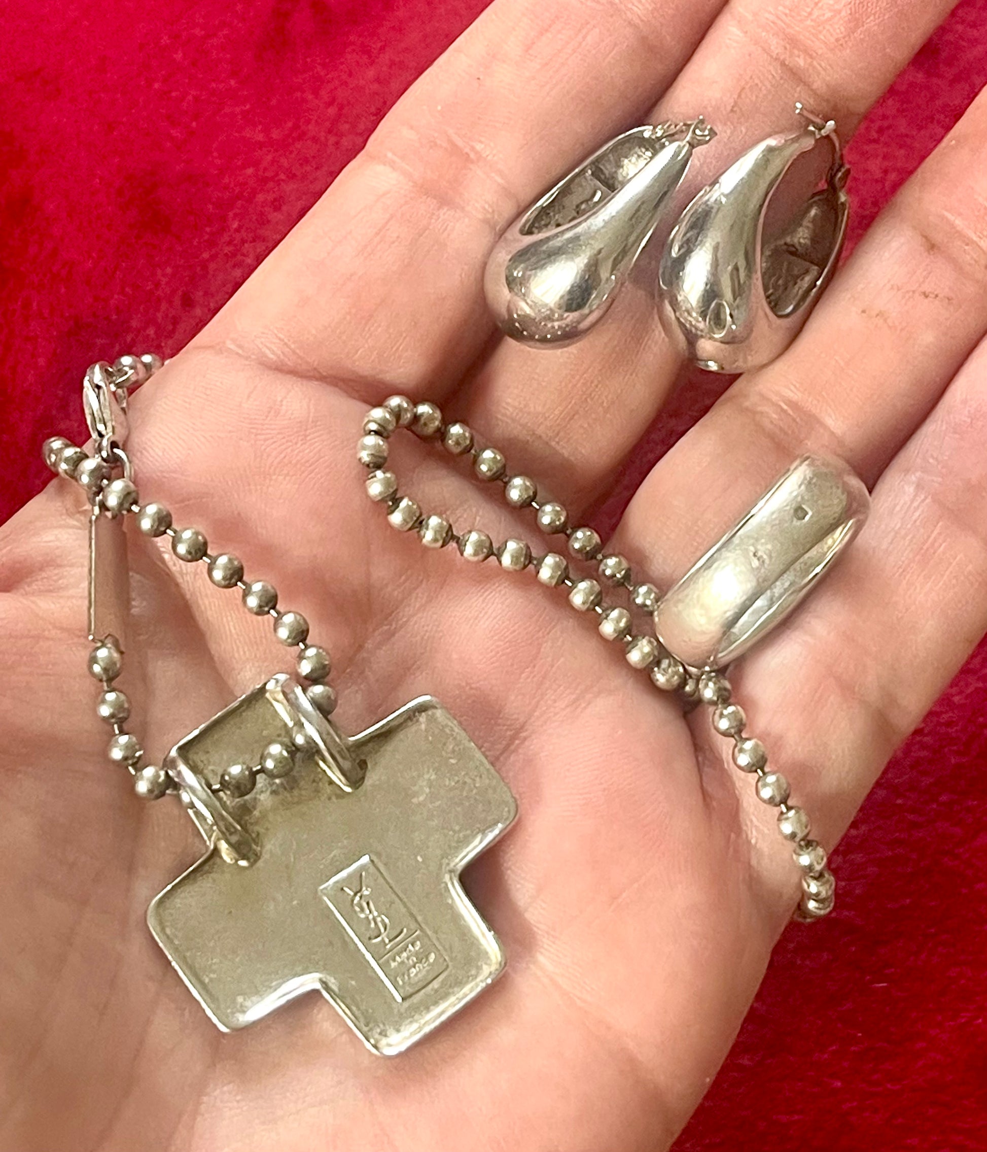Back side of Vintage Yves Saint Laurent solid silver jewelry set displayed on red fabric: silver earrings, cross pendant necklace on beaded chain, ring displayed on hand 