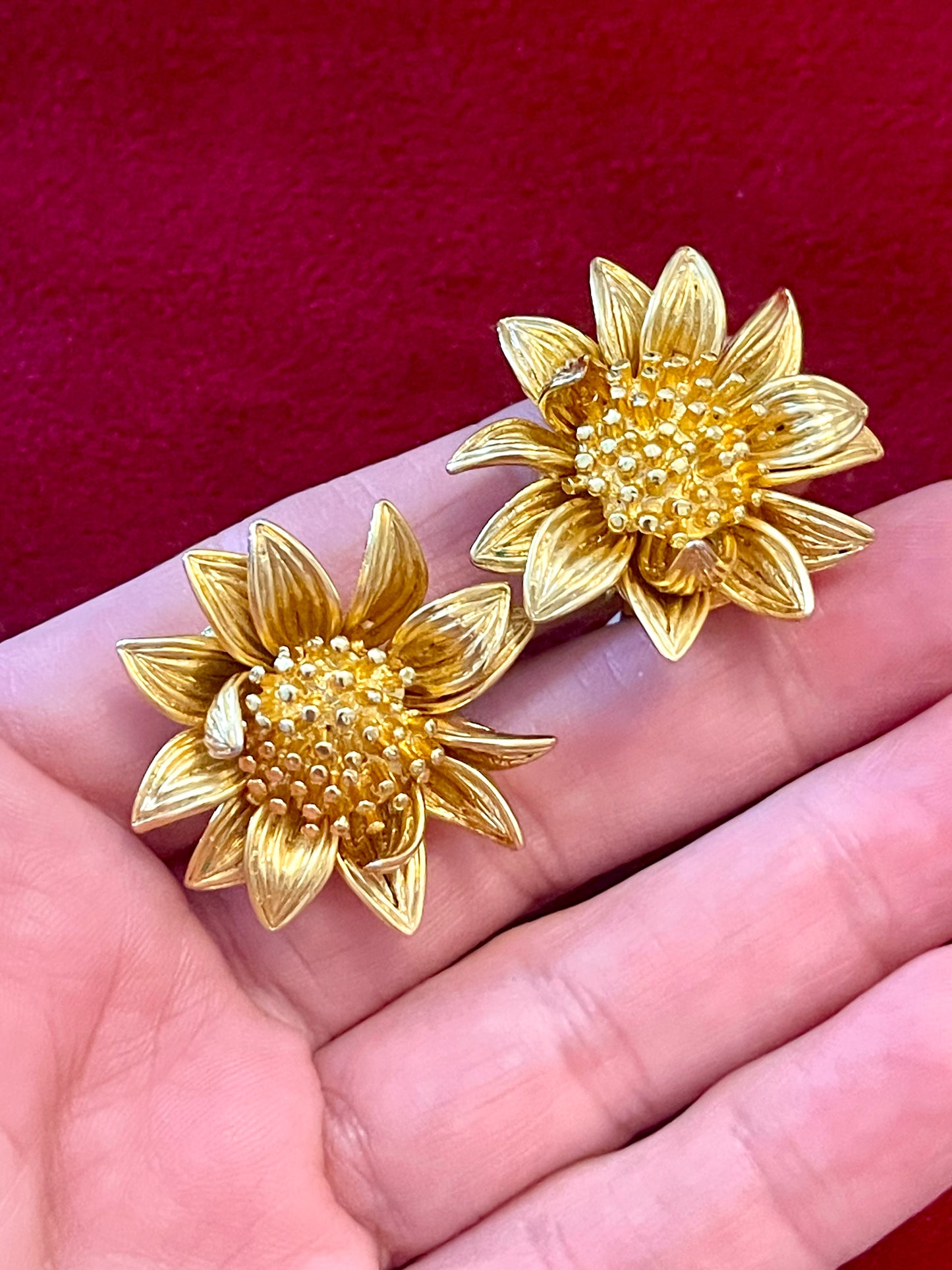Gold sunflower-shaped Orena vintage earrings held in a hand against a red background