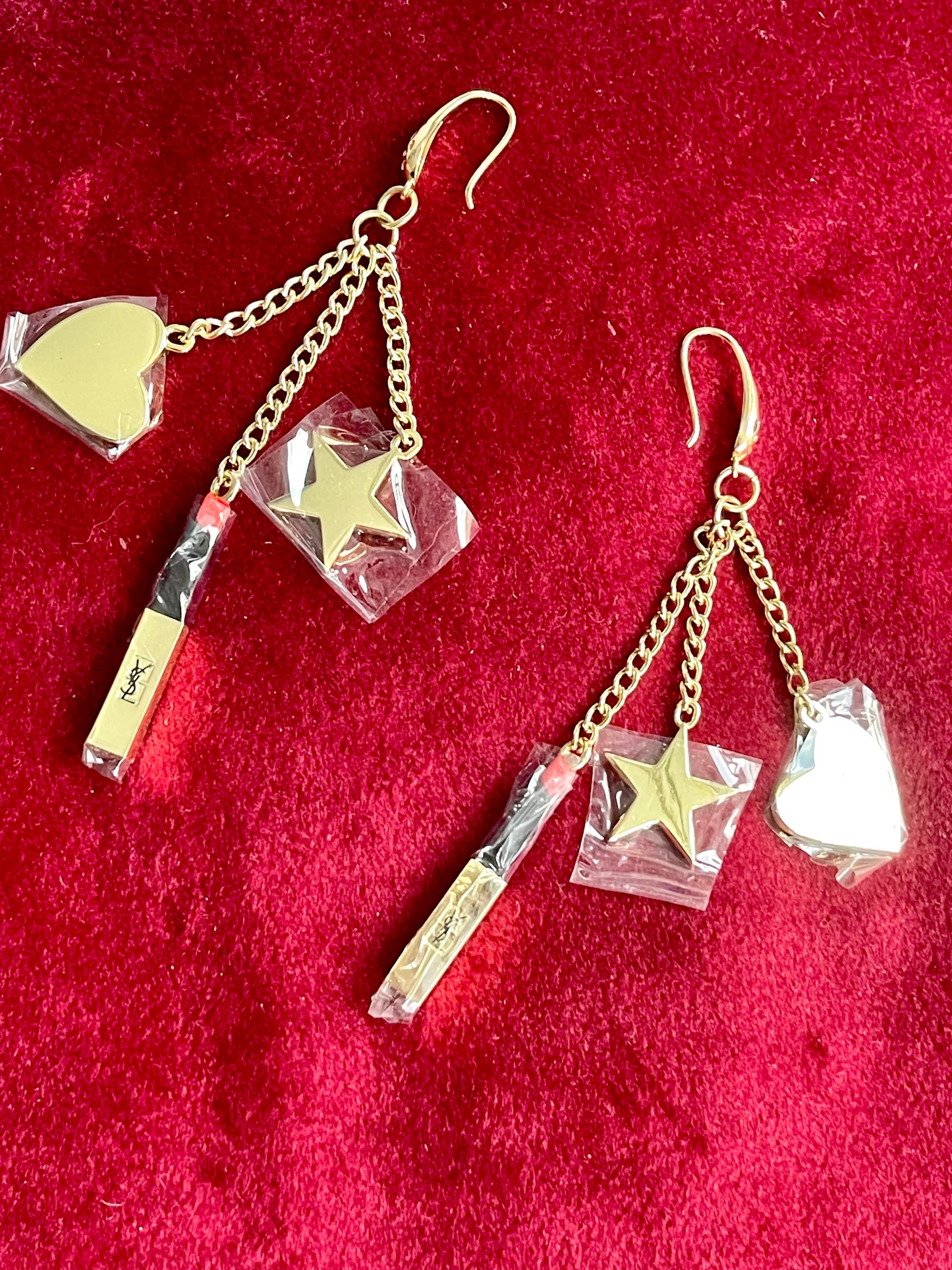 Close up view of Front view of Yves Saint Laurent Beauté upcycled earrings with lipstick, star, and heart charms on gold-plated hoops on red background 