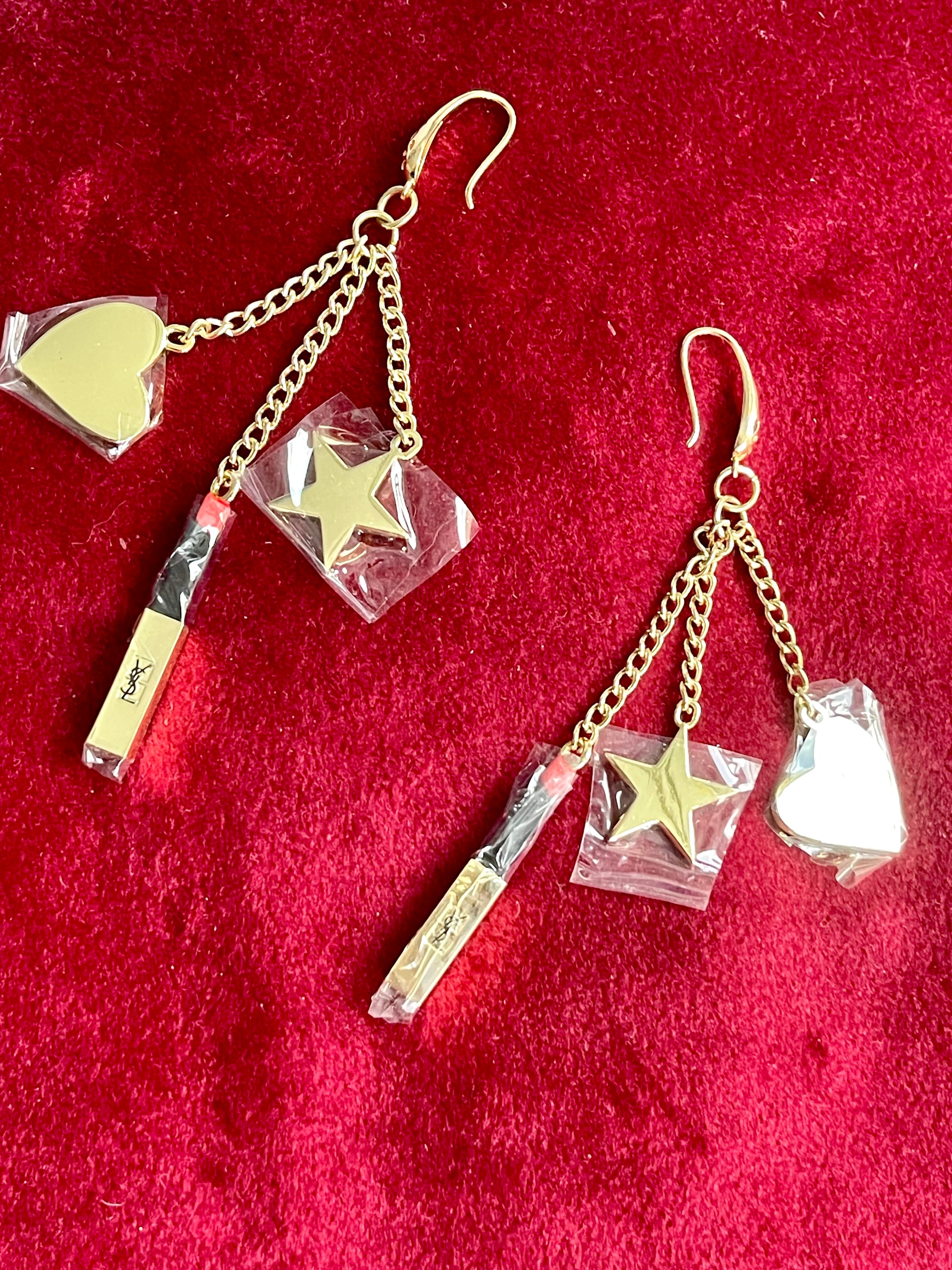 Close up view of Front view of Yves Saint Laurent Beauté upcycled earrings with lipstick, star, and heart charms on gold-plated hoops on red background 