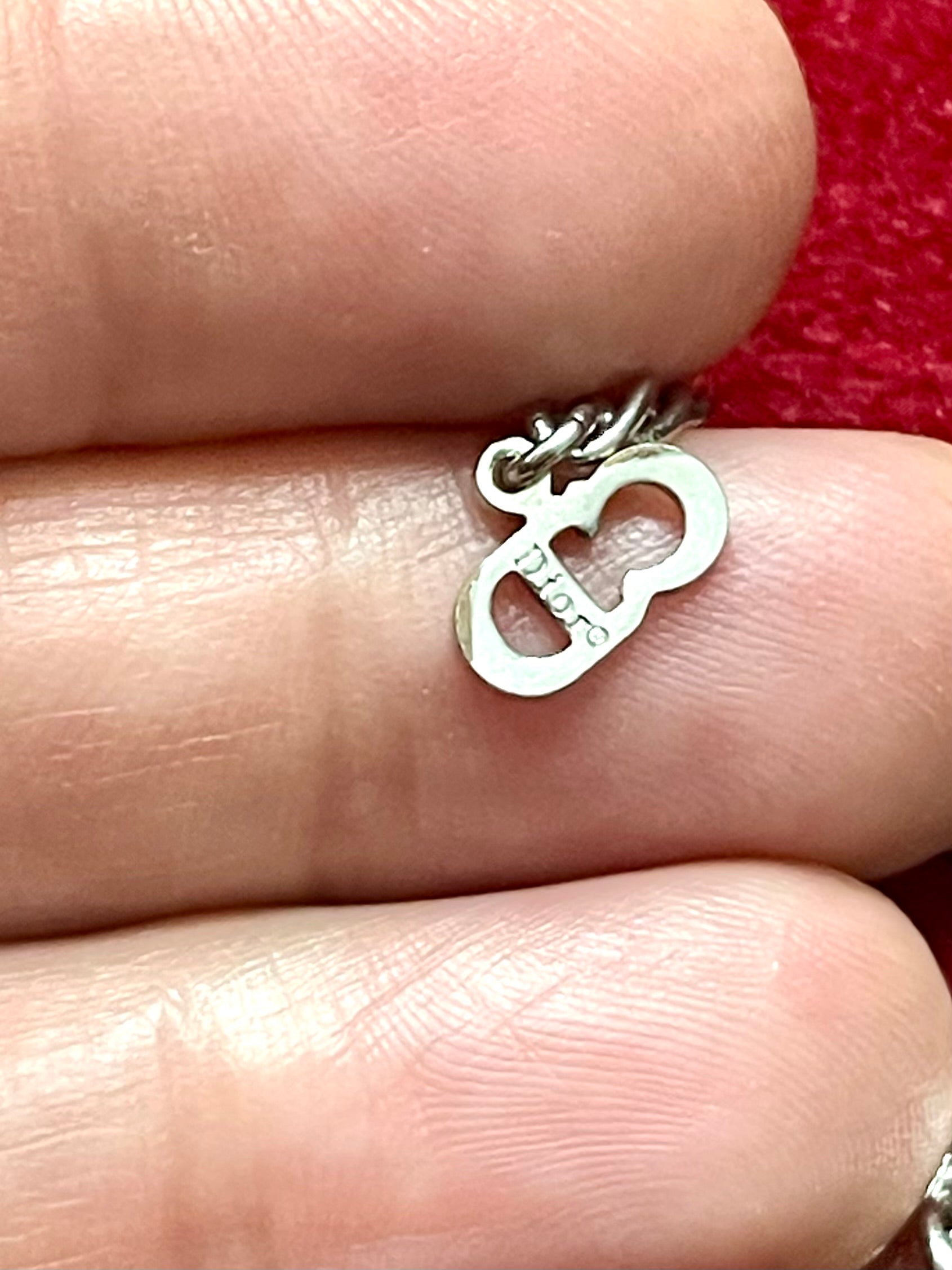 Close up on signed charm of silver Dior bracelet with a heart-shaped pendant held between fingers against a red fabric background
