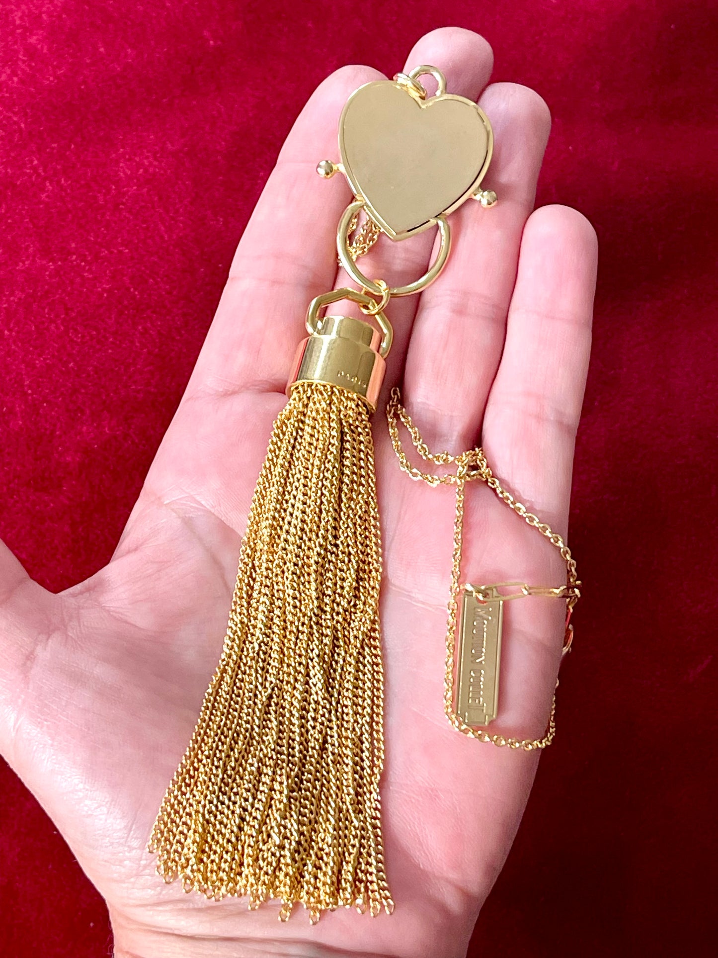 Back view on hand of Gold-plated necklace from Mutton Collet with YSL tassel charm and branded chain tag shown on red velvet fabric