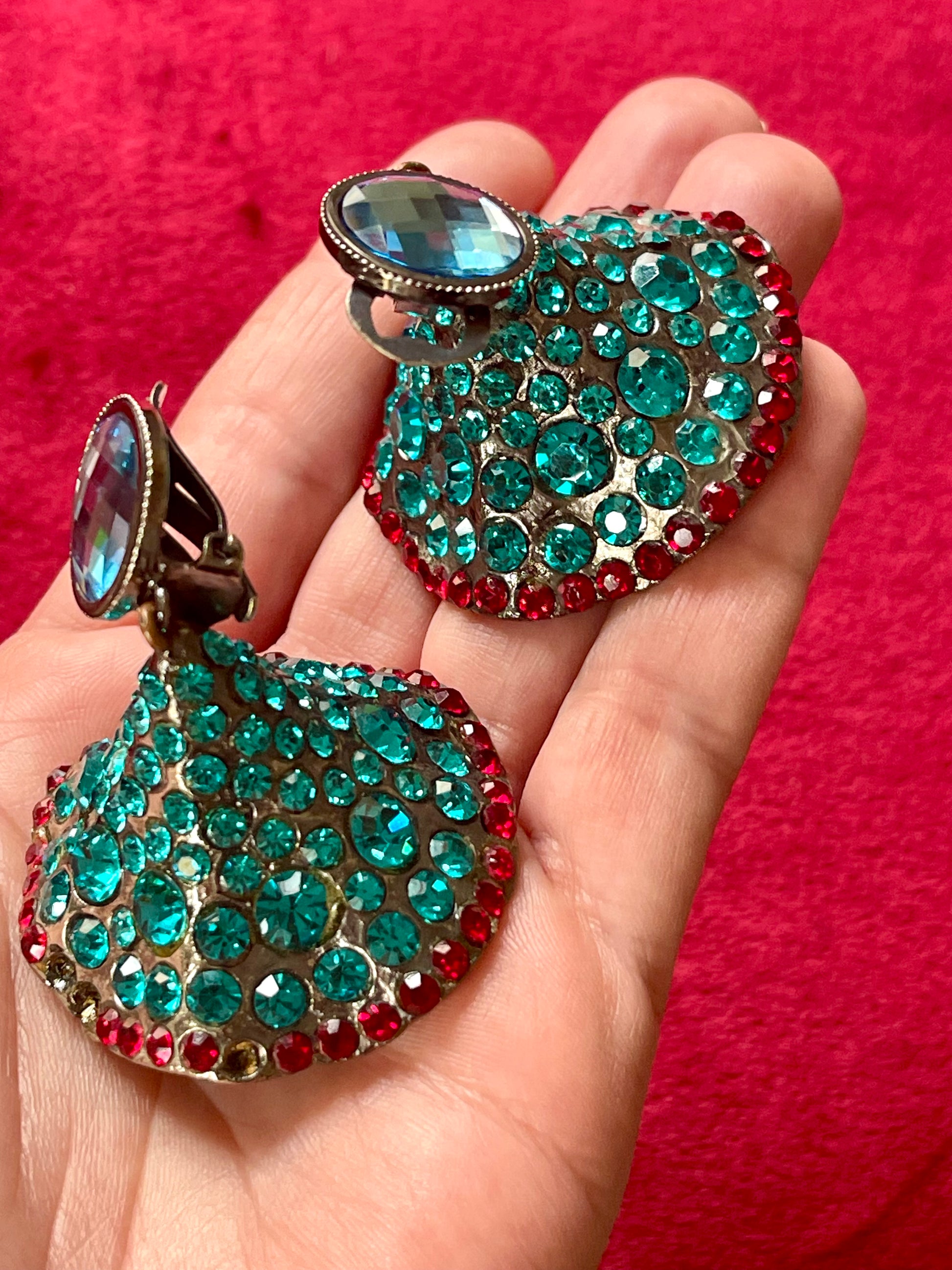 Close-up of Yves Saint Laurent Rive Gauche vintage statement earrings with turquoise and red rhinestones held in hand against a red velvet background.