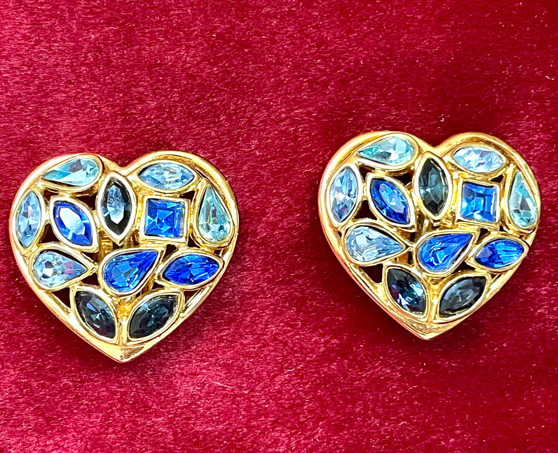 Pair of 3 cm gold-tone vintage Yves Saint Laurent heart-shaped clip-on earrings decorated with multi-coloured crystals, displayed on a red background