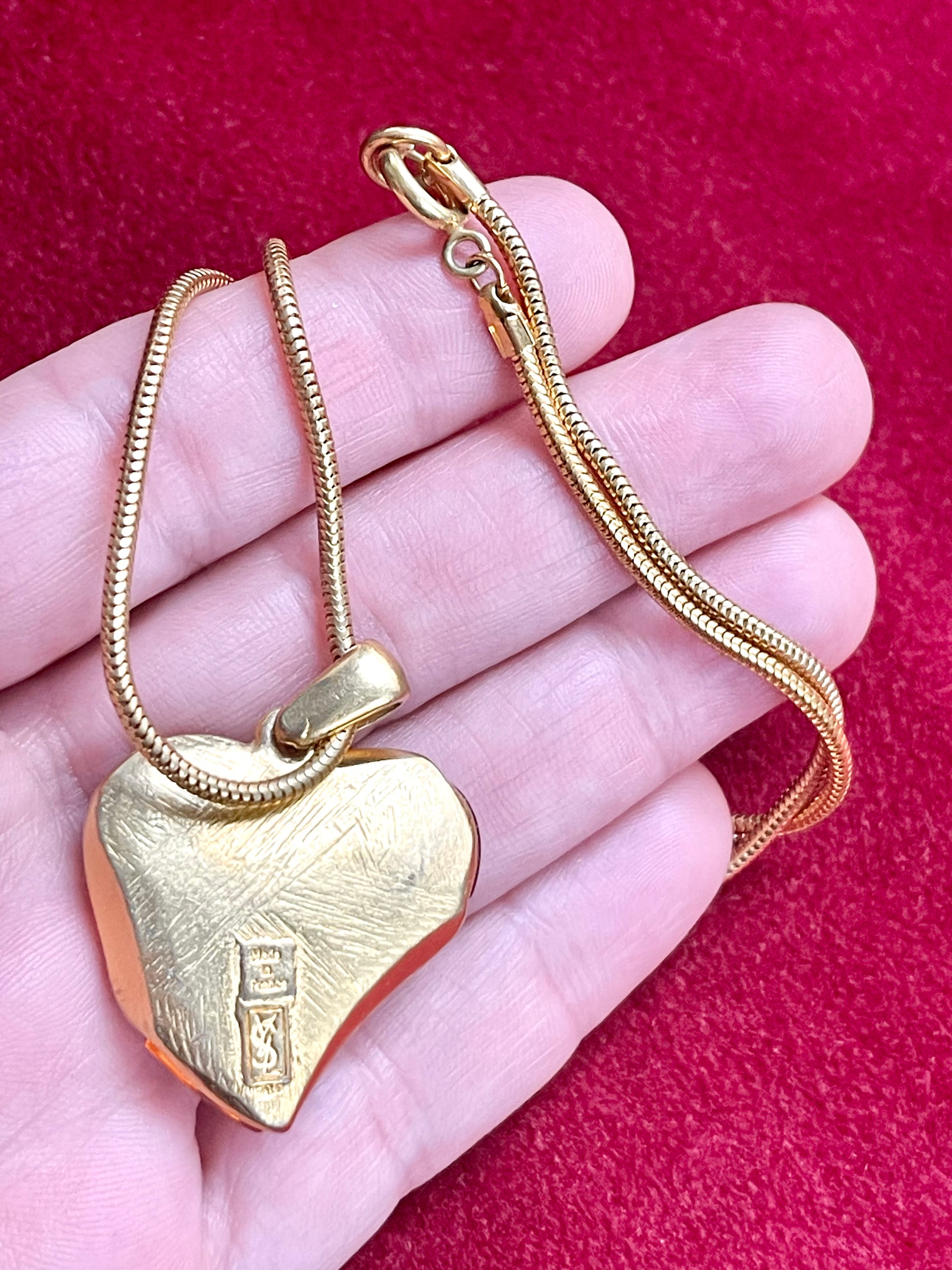 Gold heart-shaped YSL pendant on a chain held in a hand against a red background showing backside signature 