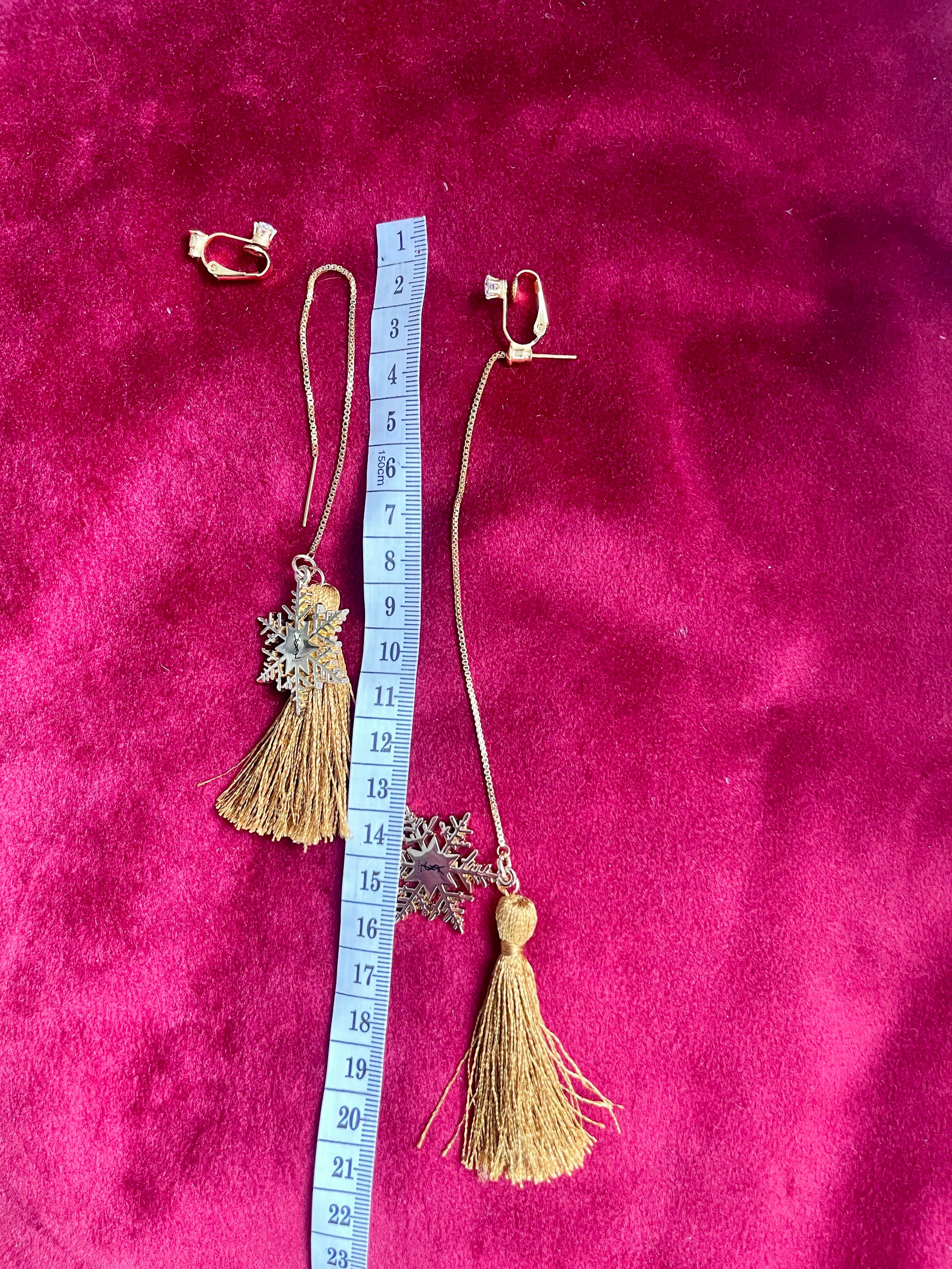 Gold earrings with tassel and snowflake pendant on a pink textured background with a measuring tape for scale.