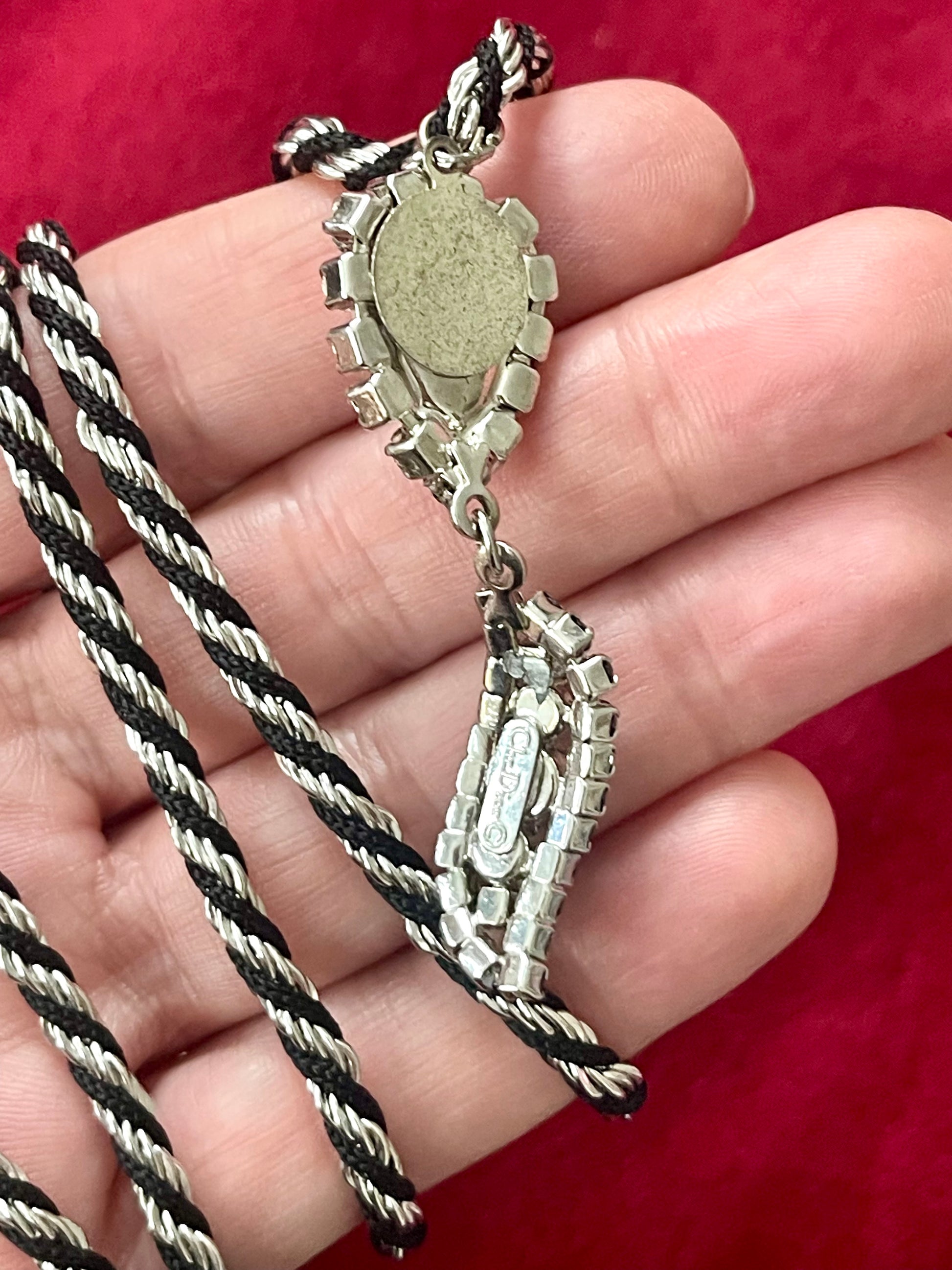 Backside view of Vintage Trifari silver-tone rope necklace with black cord featuring an attached Christian Dior crystal pendant, photographed on hand against a red background.