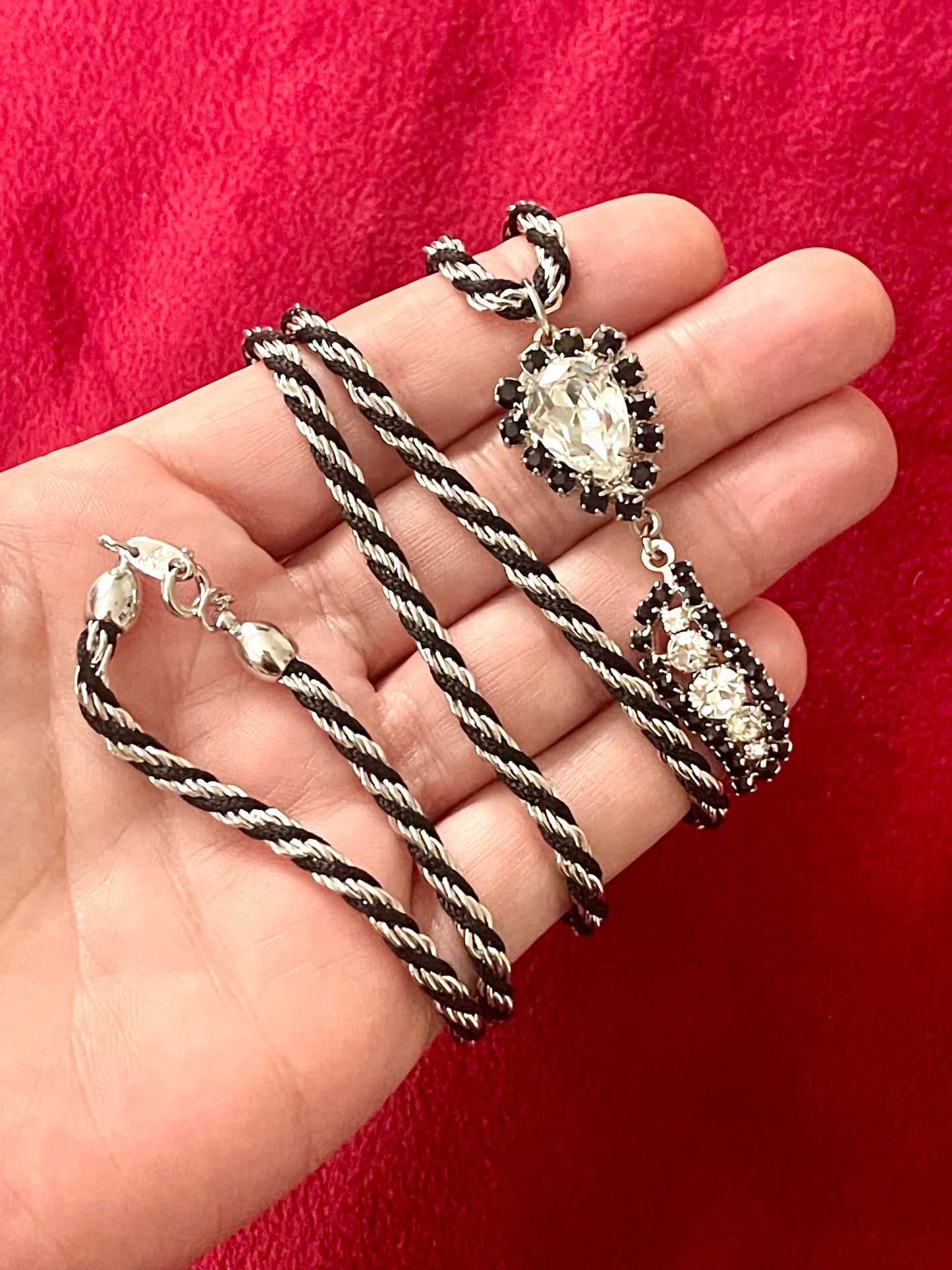 Vintage Trifari silver-tone rope necklace with black cord featuring an attached Christian Dior crystal pendant, photographed on hand against a red background.