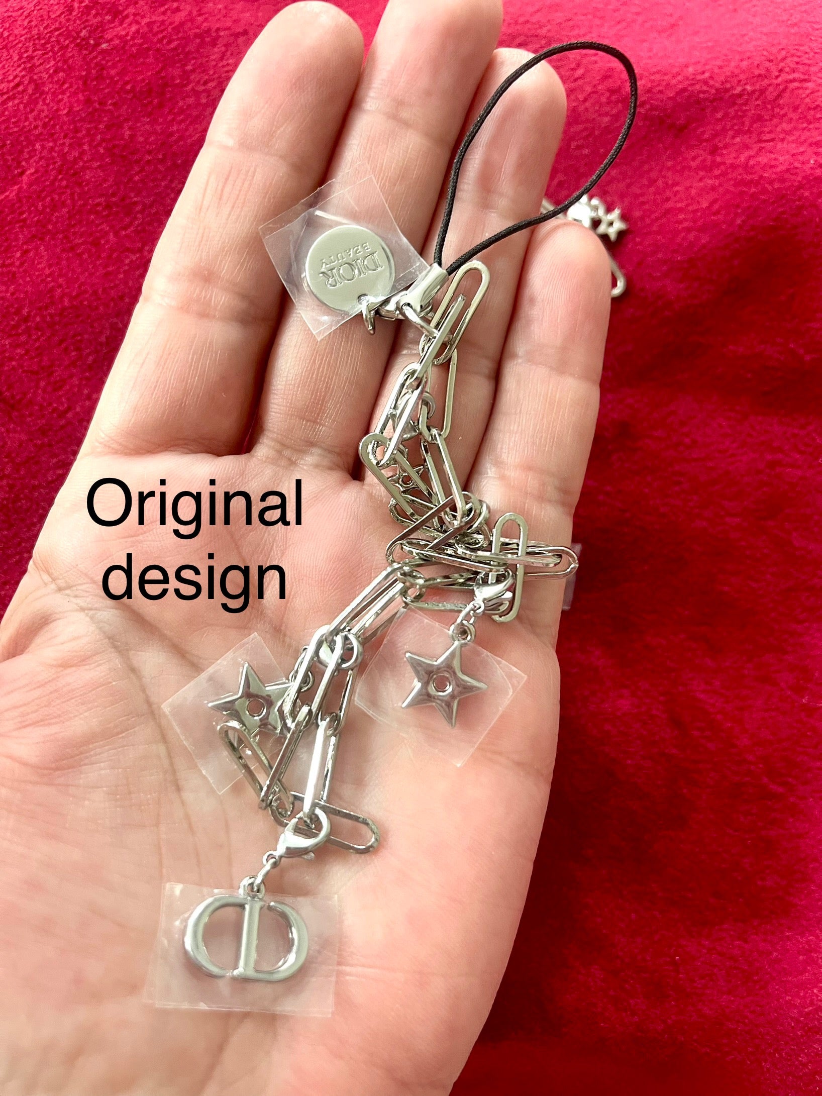Showing original design of upcycled Dior Beauté phone charm necklace with removable silver-tone charms on palm of hand with a red background 