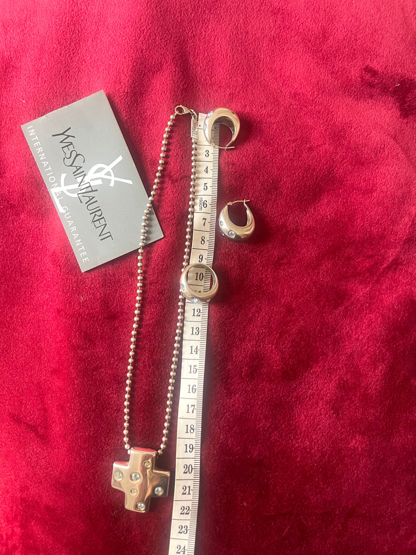 Vintage YSL silver jewelry set displayed on red fabric: silver earrings, cross pendant necklace on beaded chain, ring, measuring tape, and YSL guarantee card