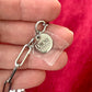 Close up on tag of upcycled Dior Beauté phone charm necklace with removable silver-tone charms held by hand with a red background 