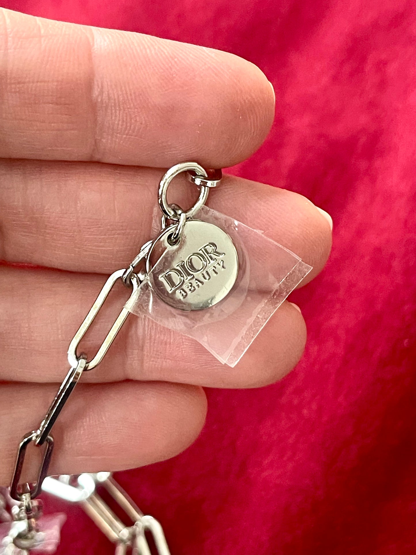 Close up on tag of upcycled Dior Beauté phone charm necklace with removable silver-tone charms held by hand with a red background 
