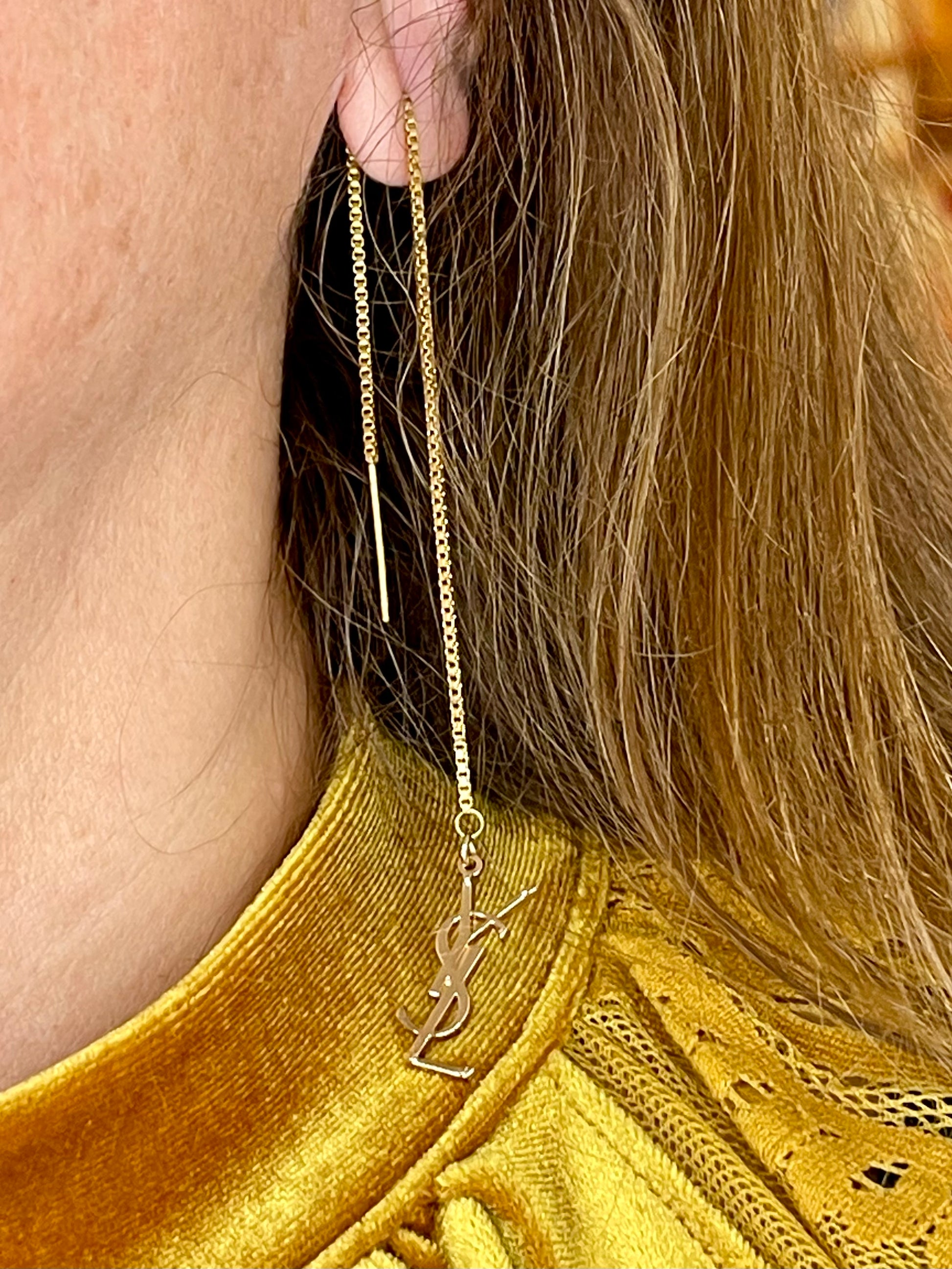Gold chain earring with a YSL logo worn by a person with long hair, wearing a mustard yellow top.
